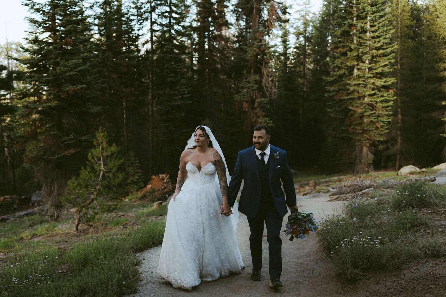 Bride and groom walk hand-in-hand on a forest path surrounded by tall pine trees in a romantic outdoor wedding.