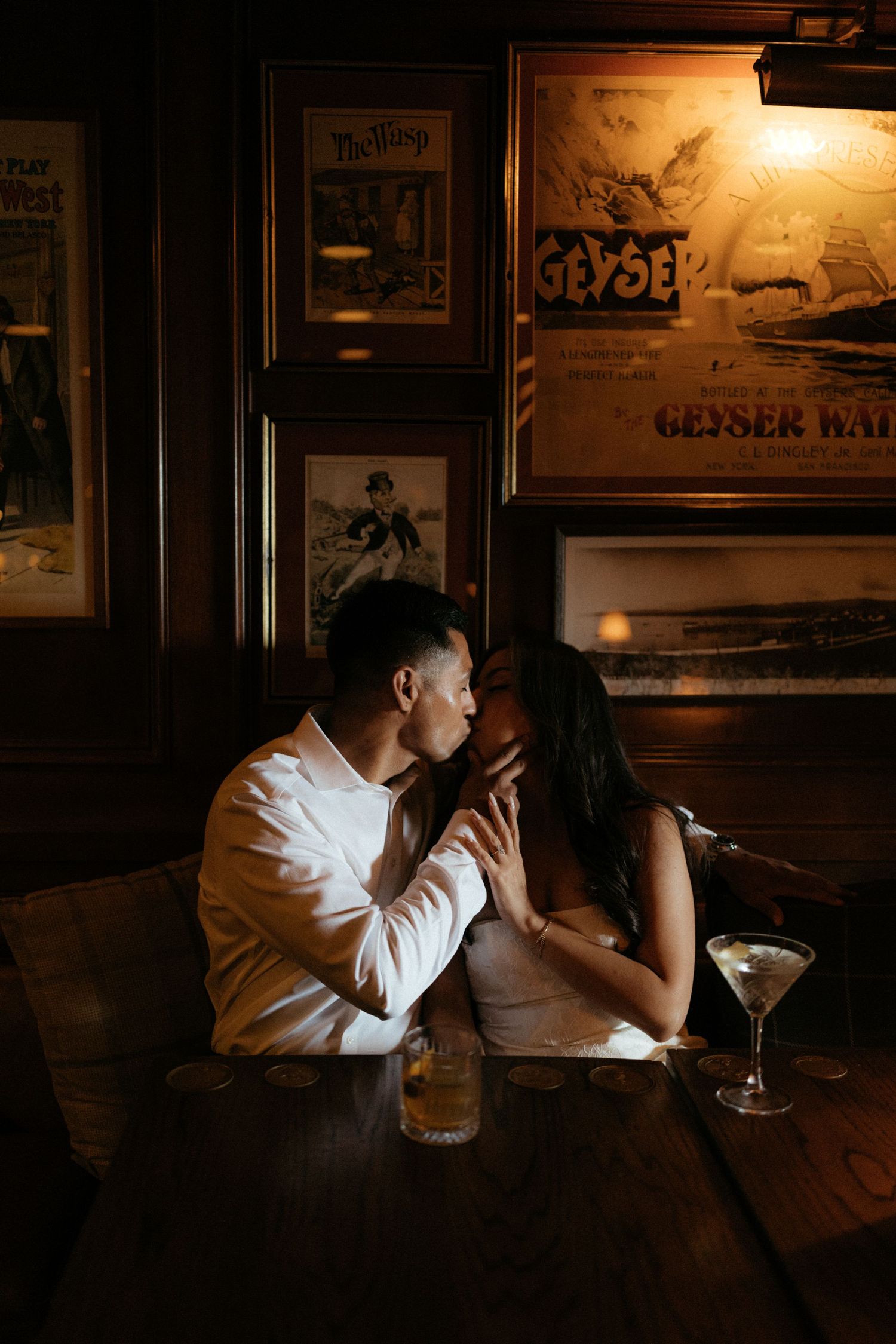 Couple sharing an intimate moment kissing at a dimly lit vintage bar with martini glasses on wooden table.