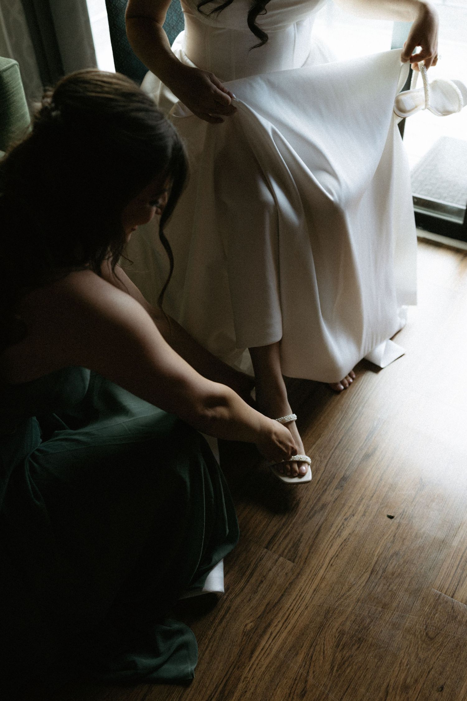 Bridesmaid in green dress helping bride put on white heels on wooden floor before wedding ceremony.
