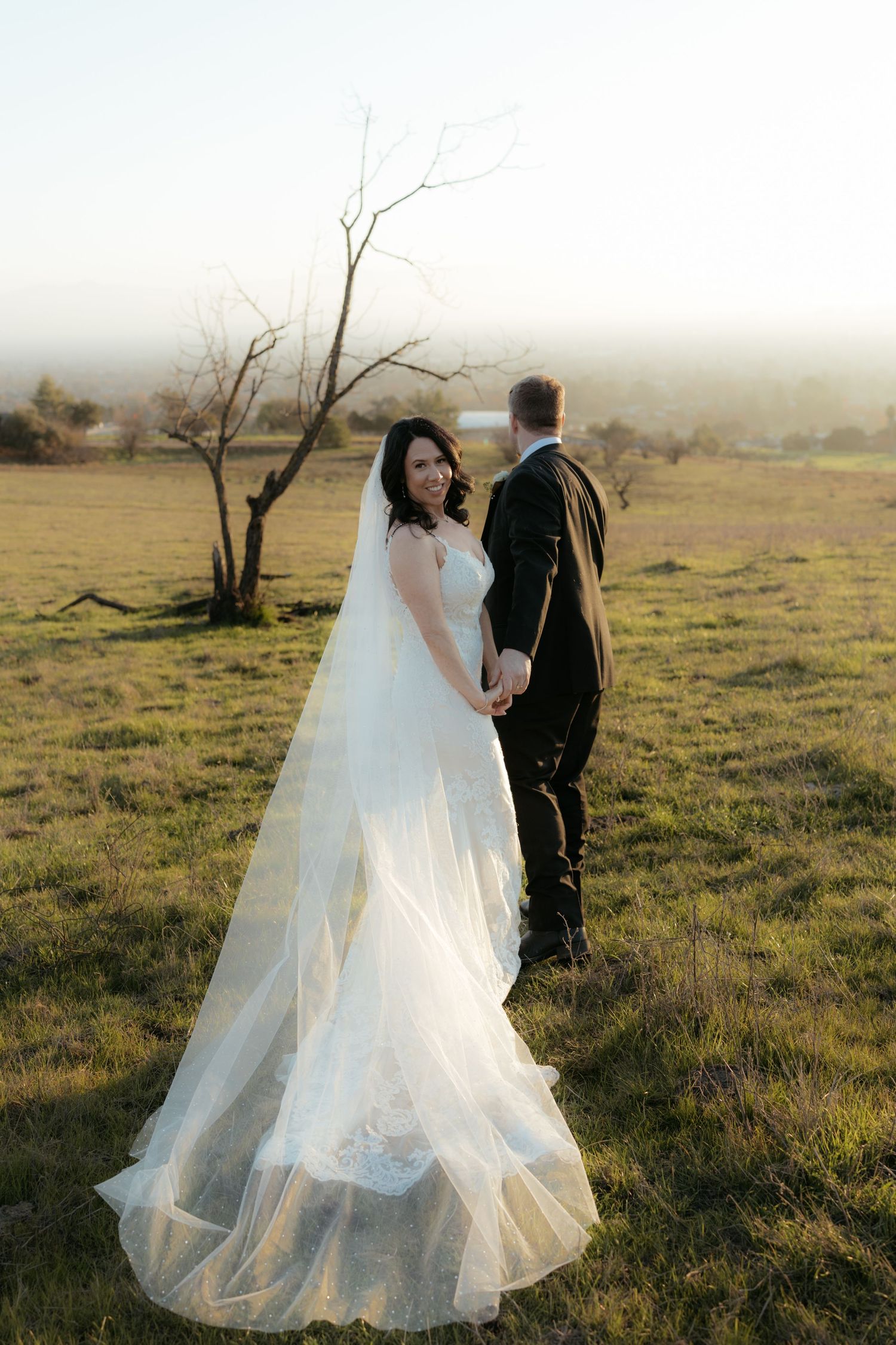 Wedding couple poses in a serene meadow with flowing white dress and dark suit.