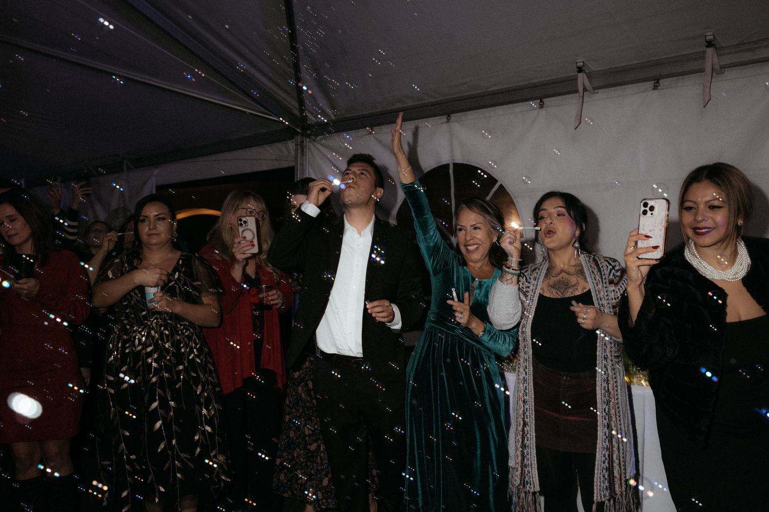 Wedding guests in formal attire celebrate on the dance floor with phones raised.