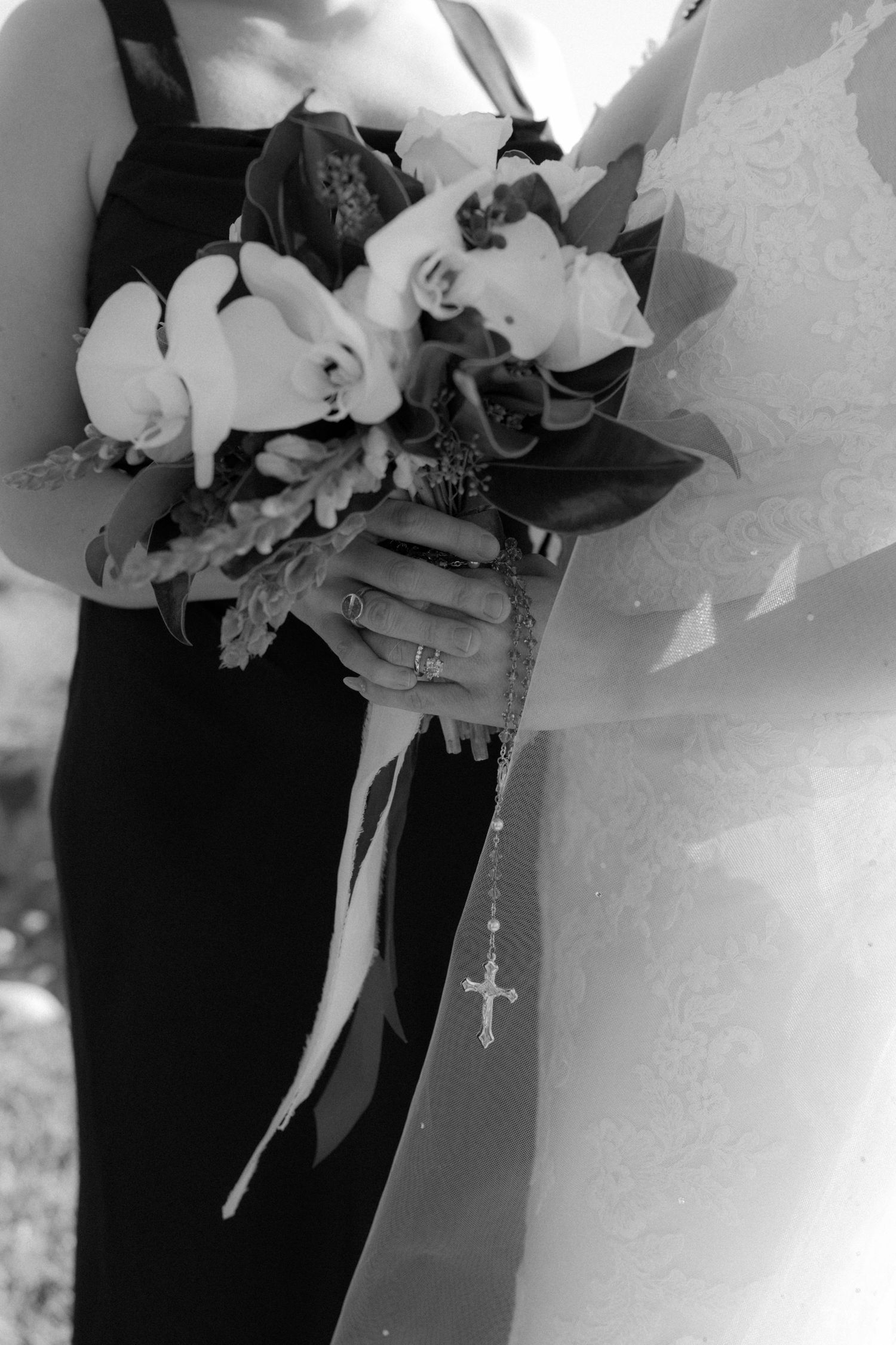 Black and white photo of a wedding bouquet with orchids and ribbons held against formal attire with a rosary visible.