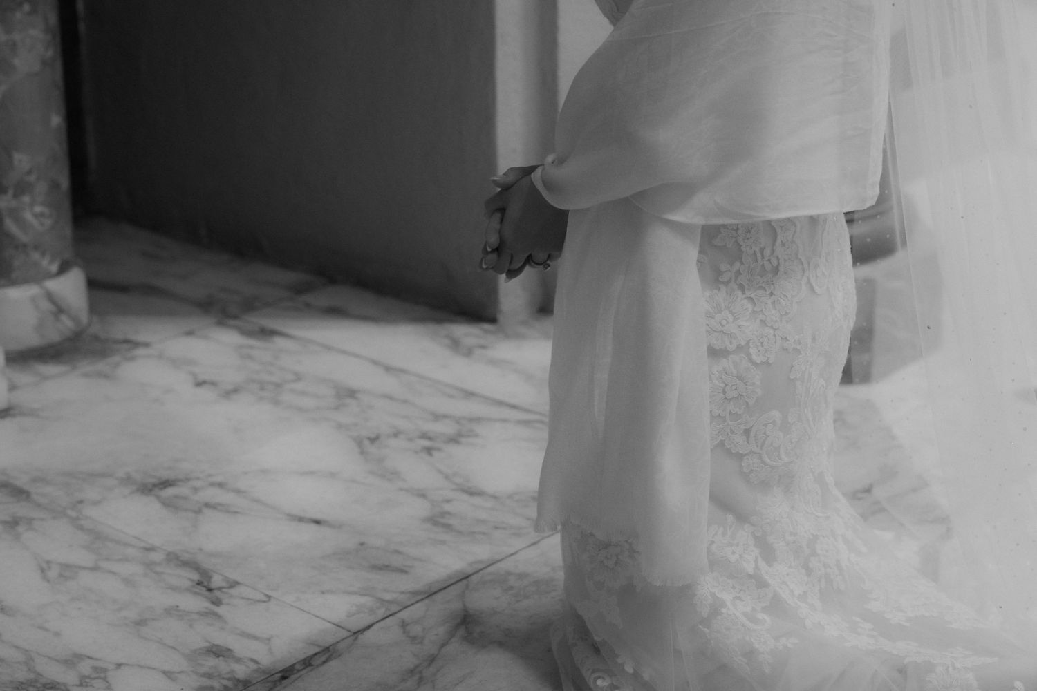 A close-up black and white detail of a bride's lace wedding dress against marble flooring.