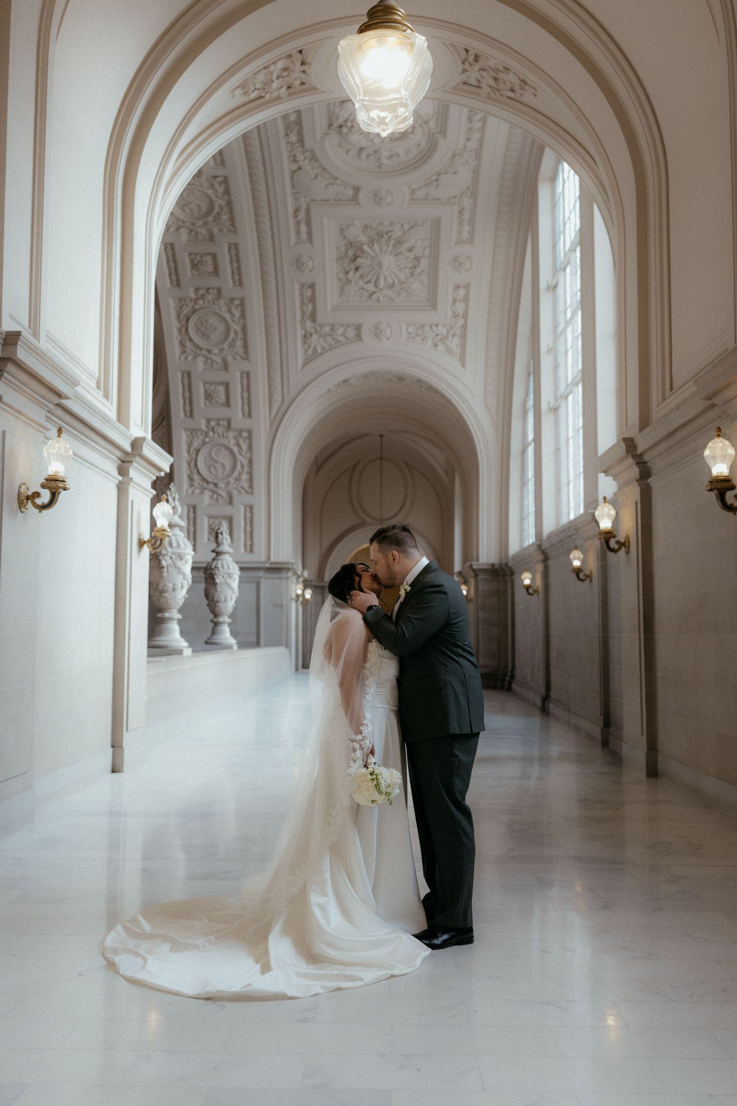 A romantic wedding couple shares intimate moments in an elegant white hallway with ornate architectural details.