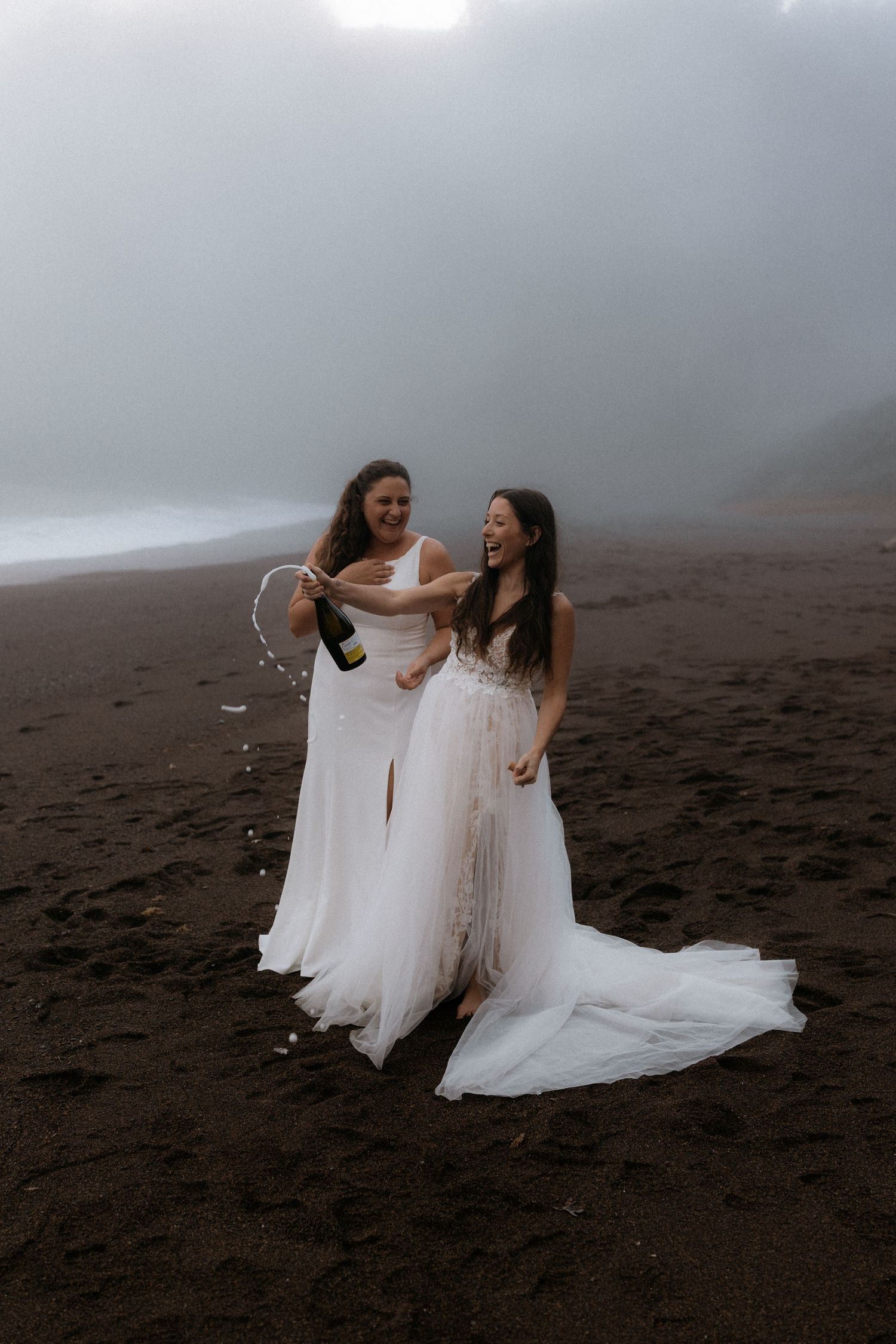 Two people in flowing white dresses dance and twirl together on a misty black sand beach.