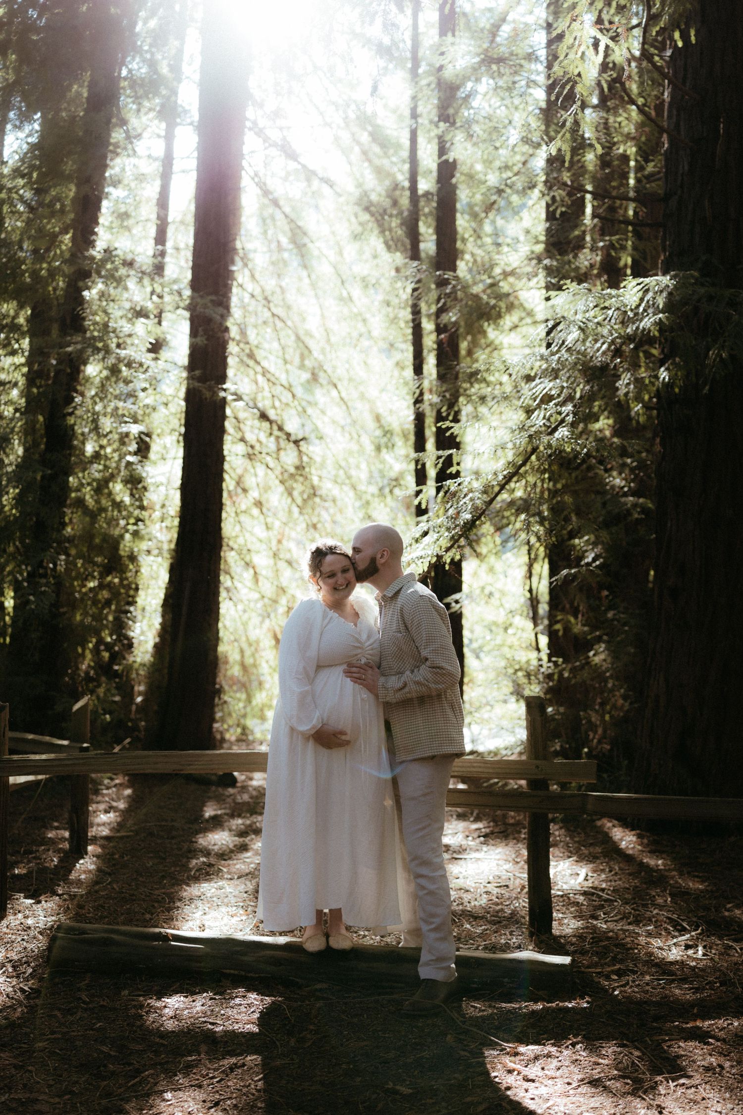 Romantic forest photoshoot with sunbeams streaming through tall trees as a couple embraces in white attire.