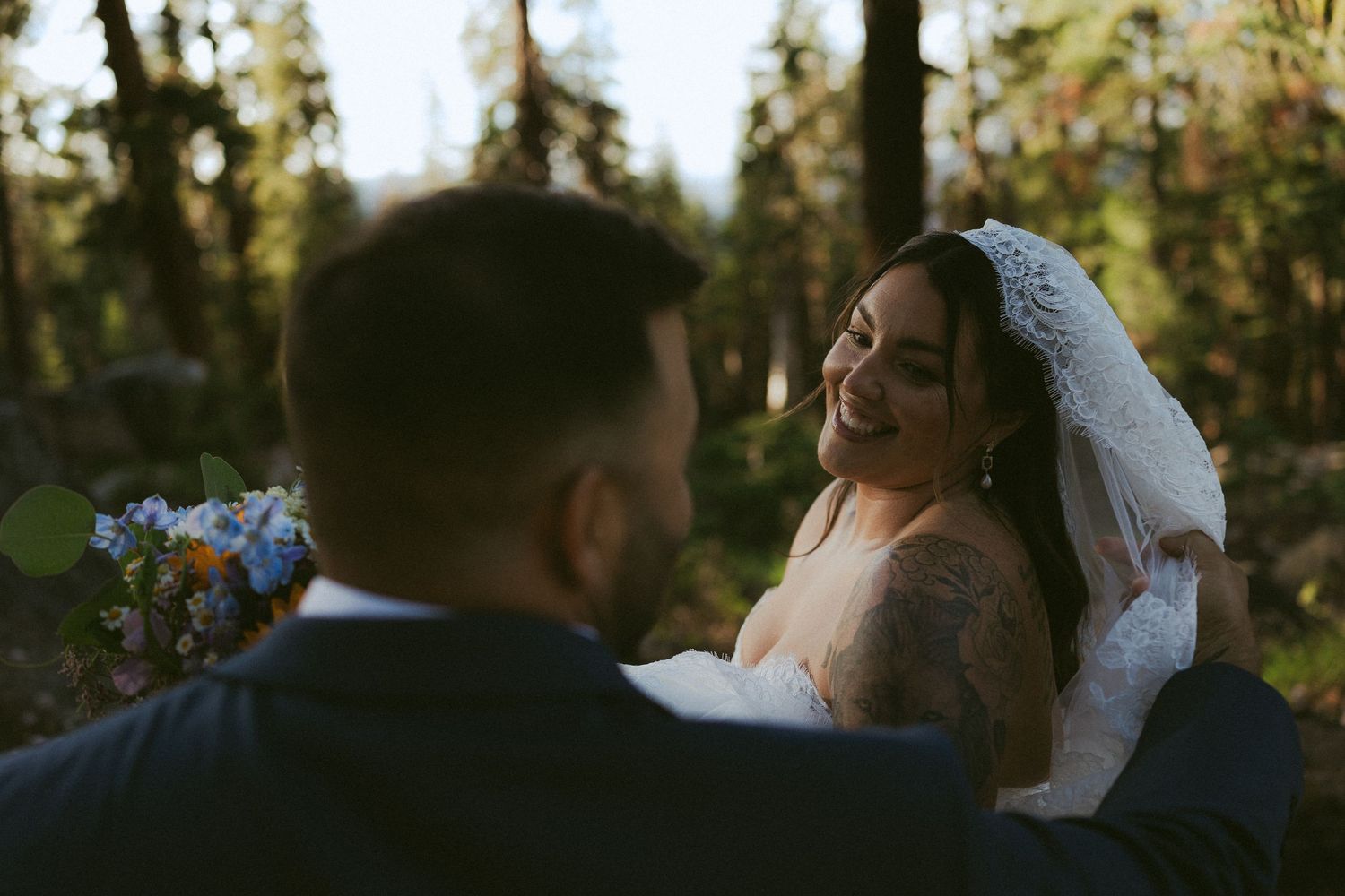 Bride in lace veil smiles at groom during outdoor forest wedding ceremony.