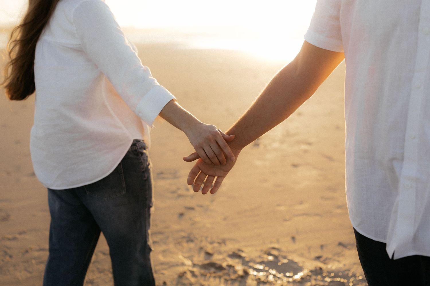 Couple holding hands on a sandy beach at sunset, wearing white shirts and jeans.