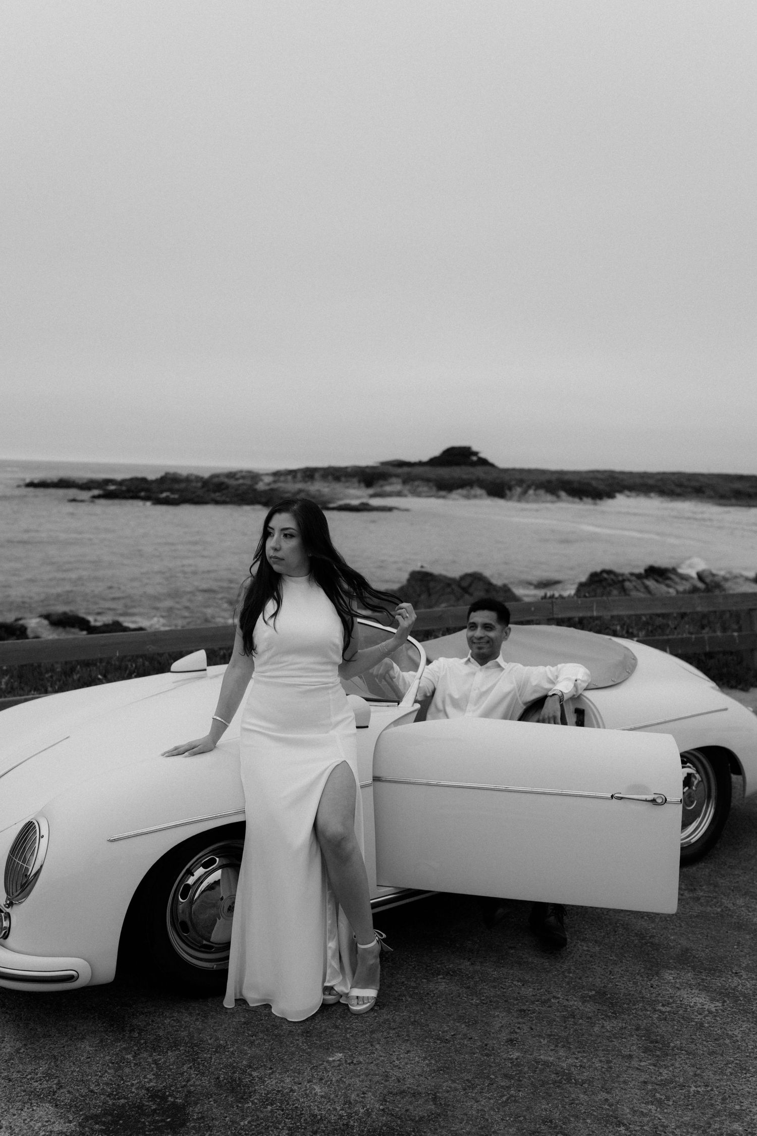 Bride in white gown poses by vintage convertible with groom seated inside, ocean backdrop, black and white photo.