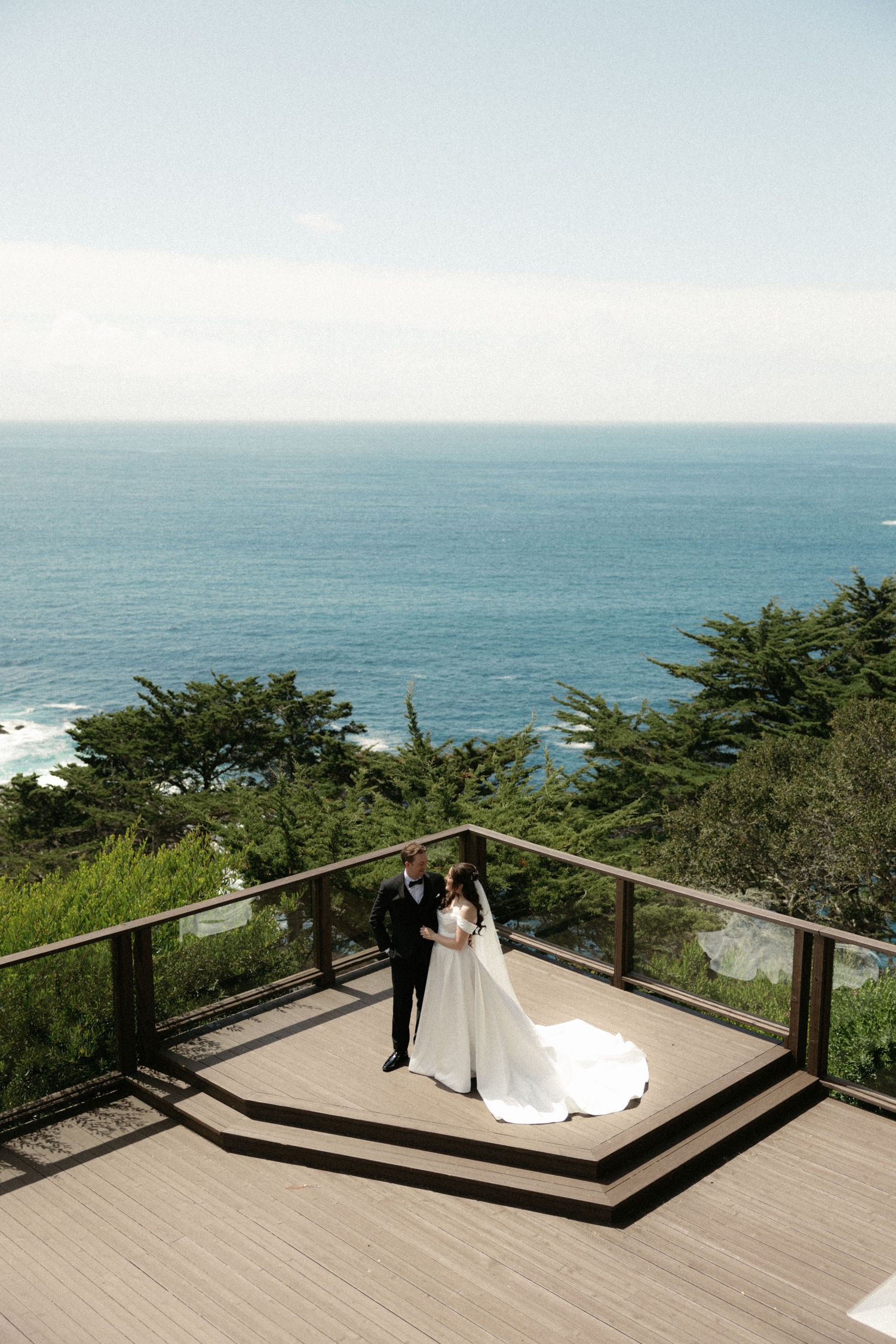 Bride and groom stand on an oceanview wooden deck, her white gown trailing, with lush trees and blue sea behind.