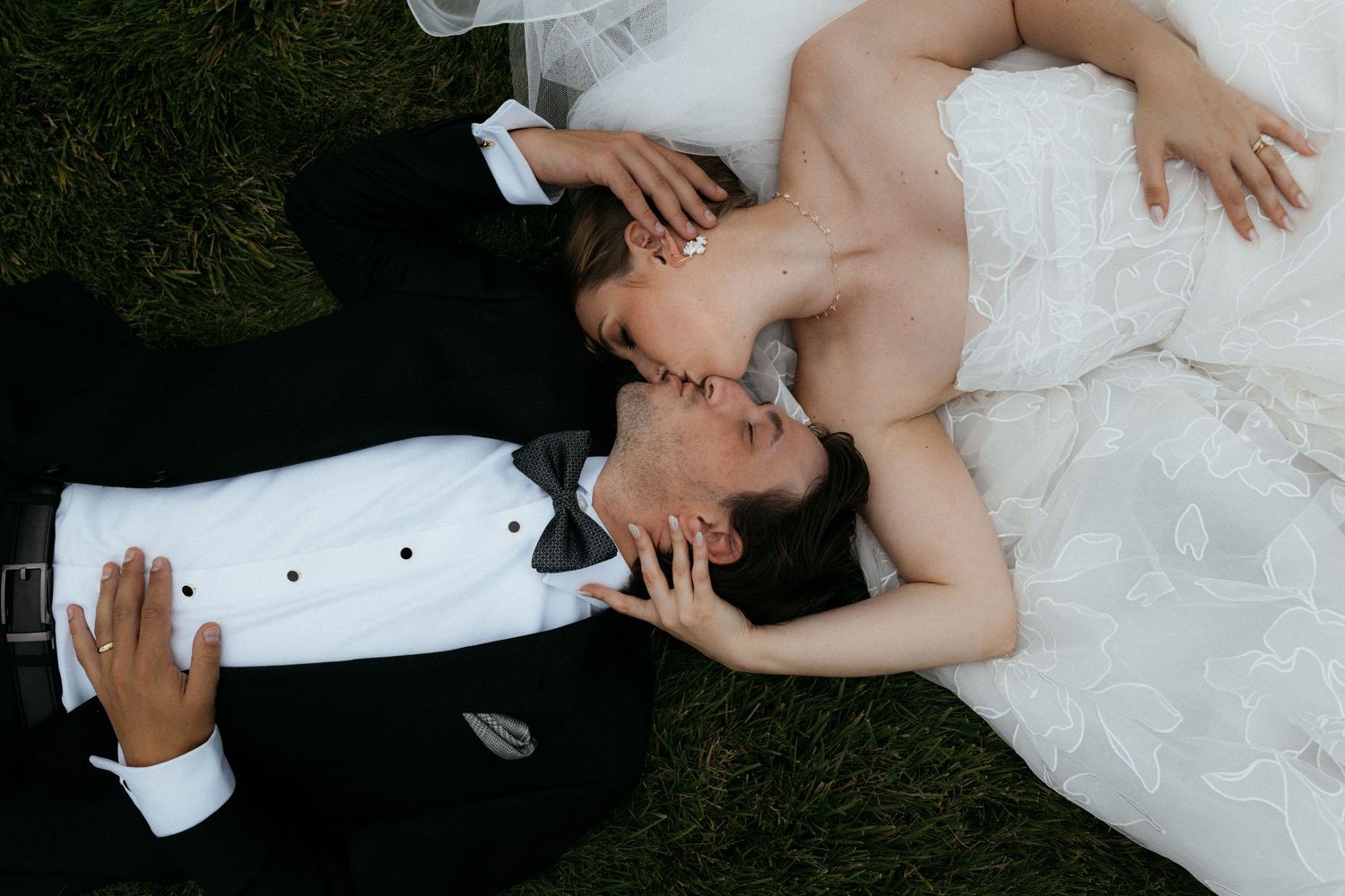 Bride and groom lying on green grass, sharing a romantic kiss in wedding attire, white gown and black tuxedo.