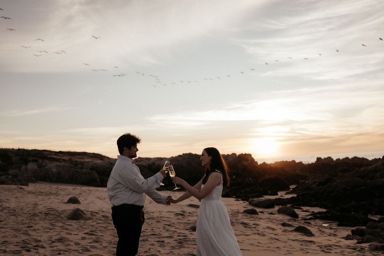 Couple walking hand in hand along rocky beach landscape at sunset.
