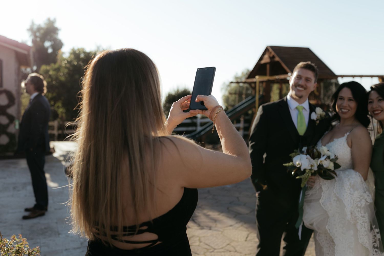 Wedding guest takes photo on smartphone of newlyweds at outdoor venue during sunset.