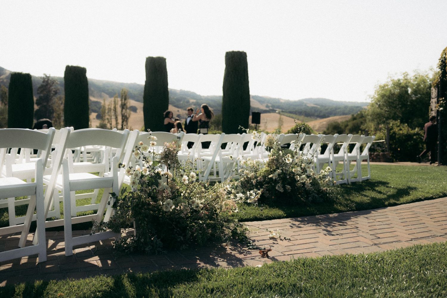 A panoramic view of a white fence along rolling green hills with mountains in the distance under a cloudy sky.