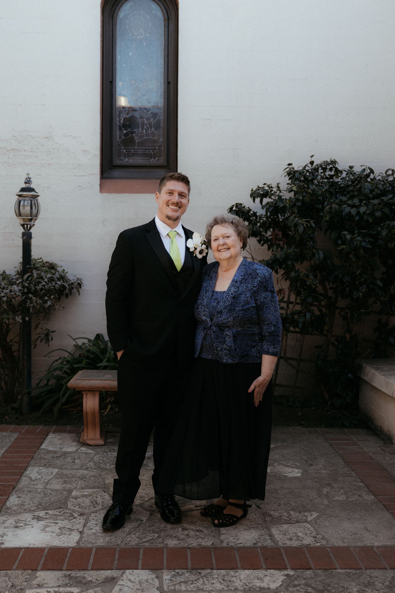 A couple in formal evening wear stands together on a stone patio beside a white stucco wall and garden.