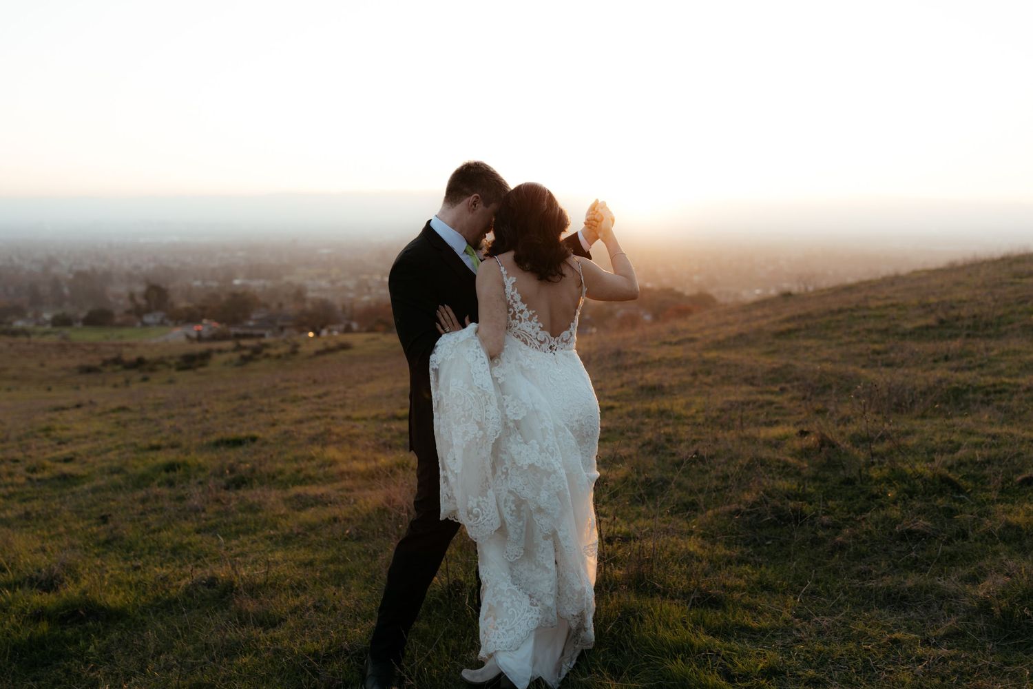 A couple in wedding attire embraces on a misty hilltop during sunset while a photographer captures the moment.