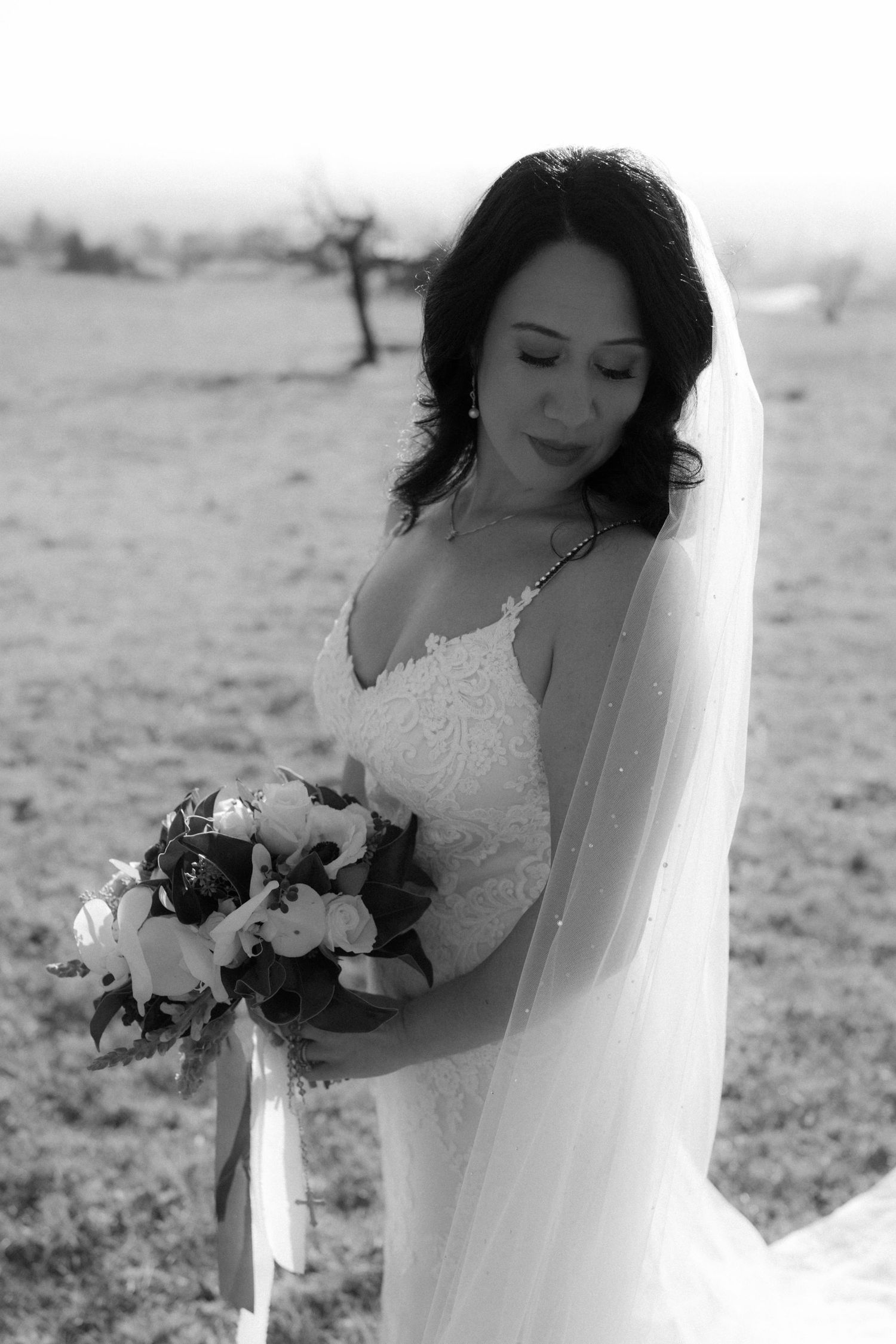 Black and white portrait of a bride in a lace gown holding a floral bouquet in a desert landscape.