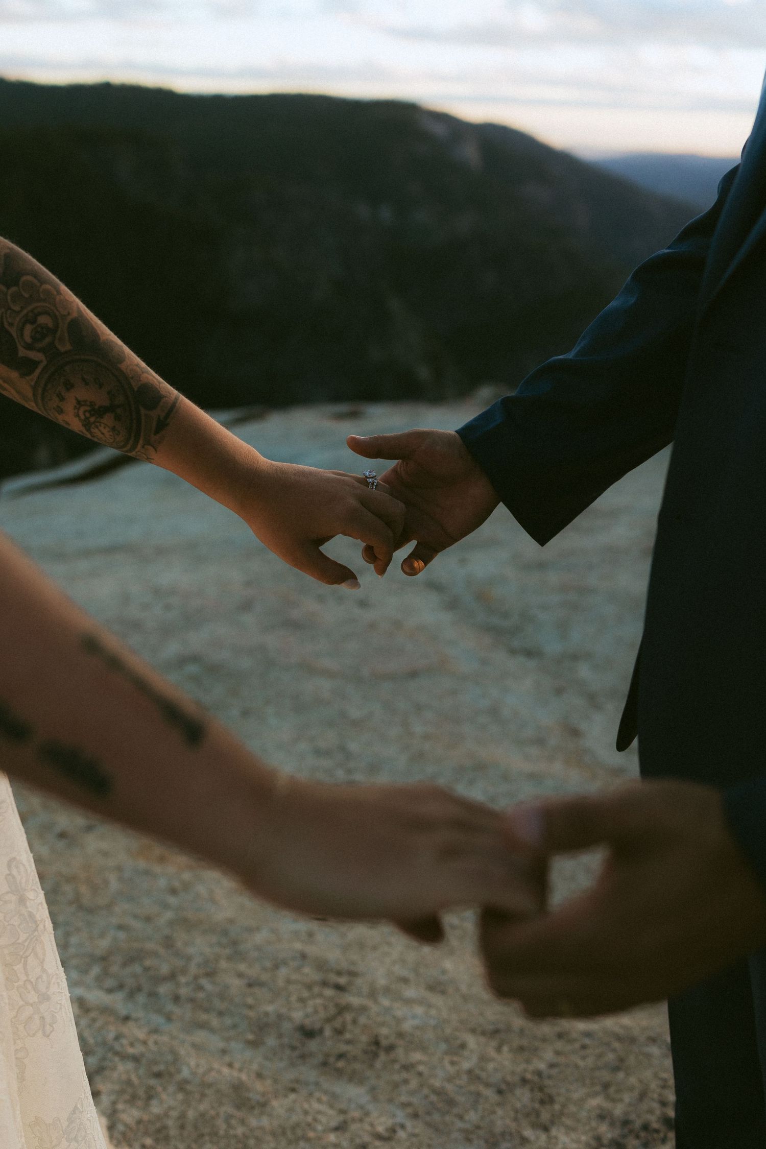 Couple holding hands with tattooed arm and wedding ring on rocky mountain overlook at sunset.