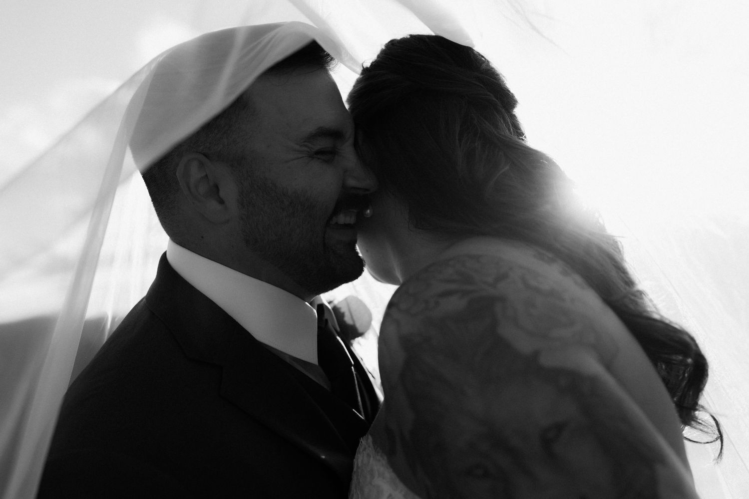 Black and white wedding photo of bride and groom sharing a romantic kiss under a flowing veil.