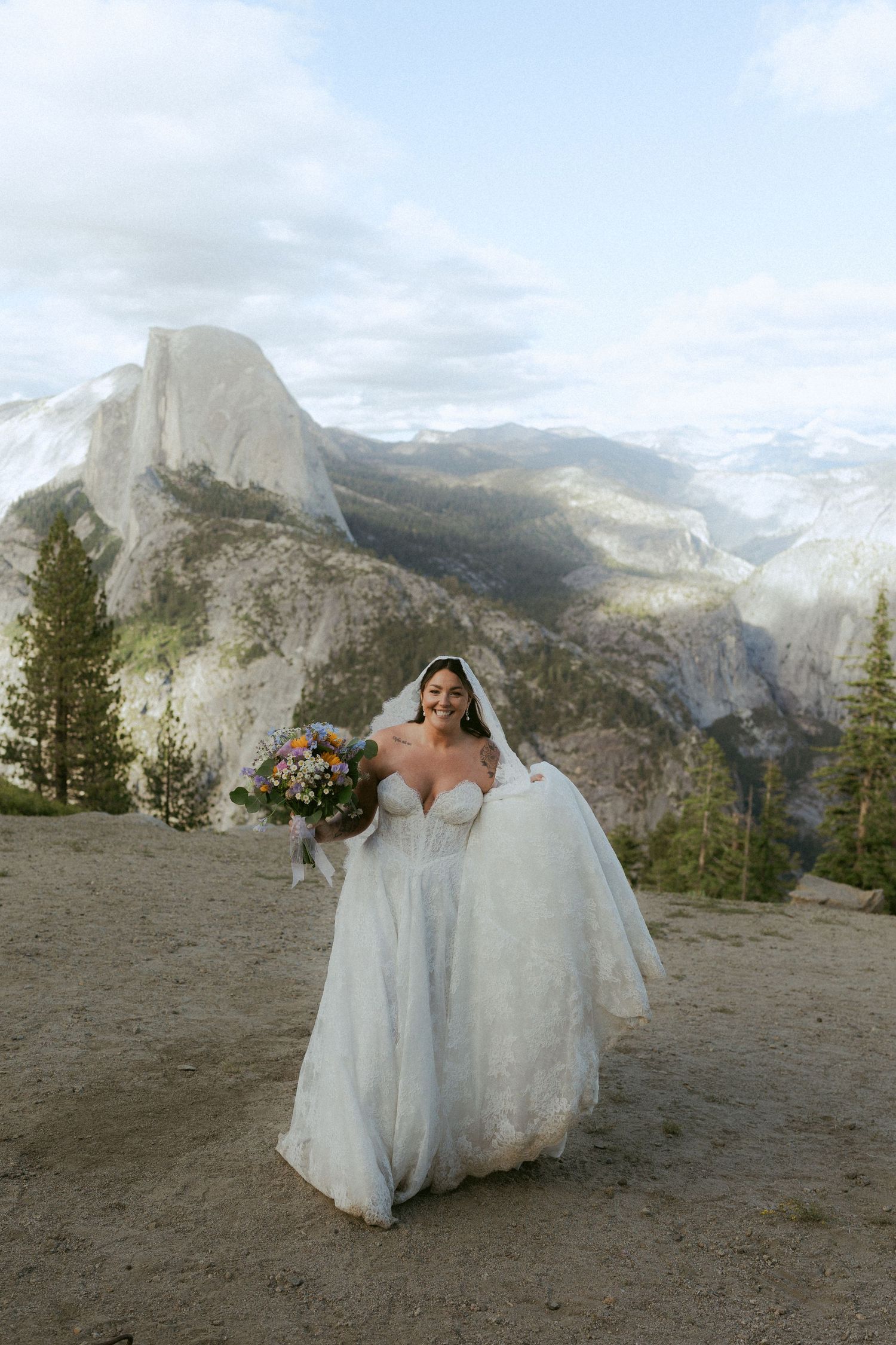 Bride in white gown holding colorful bouquet smiling at Glacier Point with Half Dome in background at Yosemite.