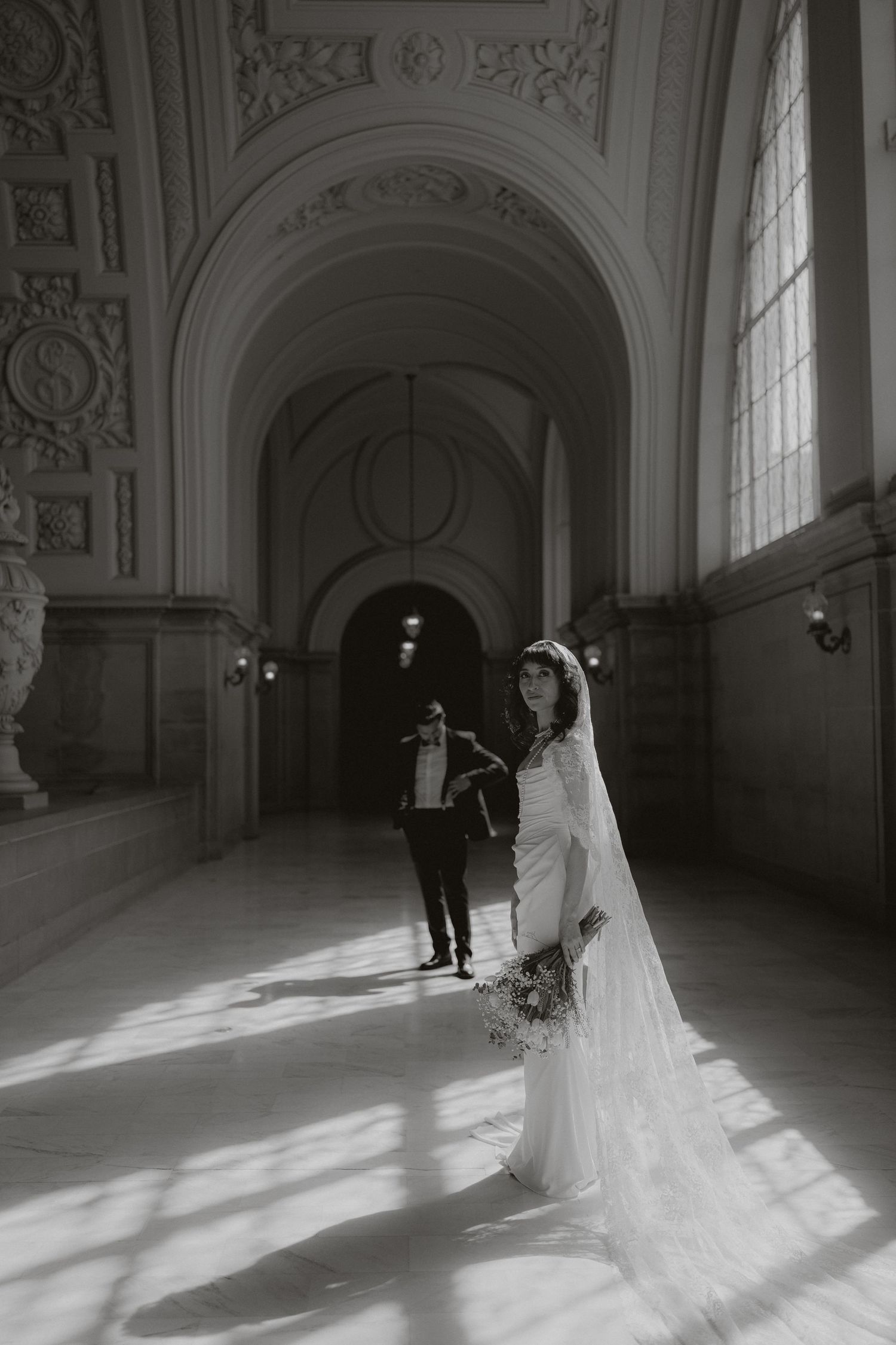 A bride in a flowing white dress and veil stands in an ornate hallway as light streams through tall windows.