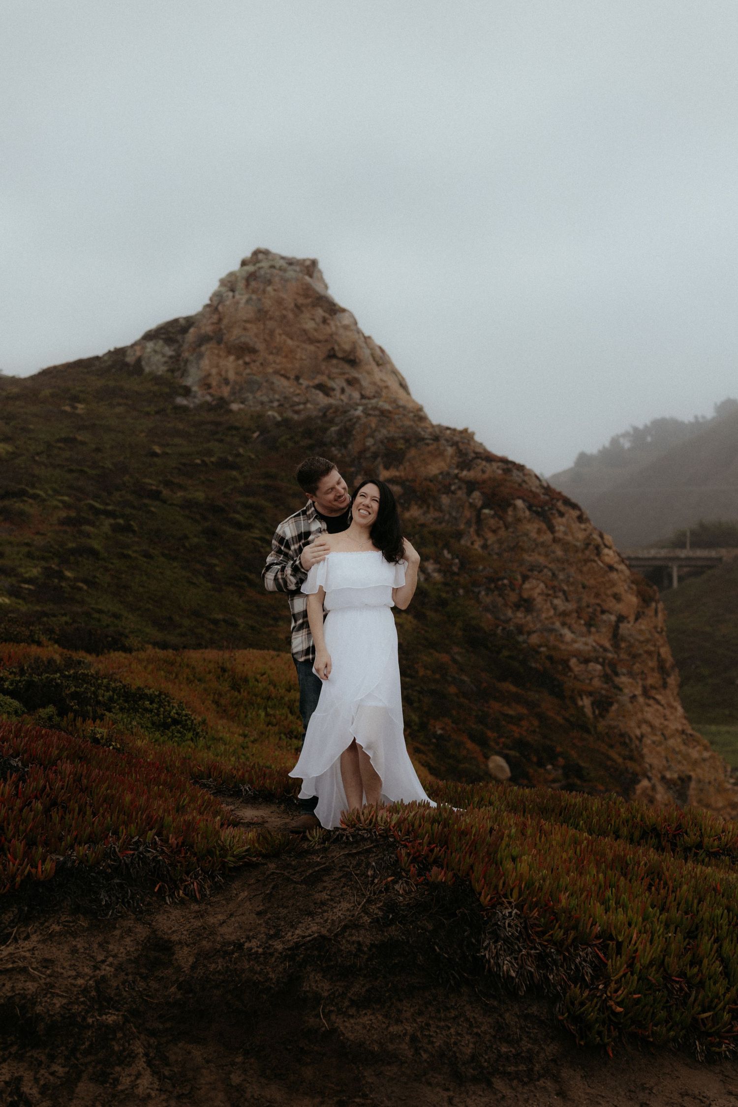 A romantic photoshoot on a misty mountainside with dramatic rocky cliffs and coastal vegetation.