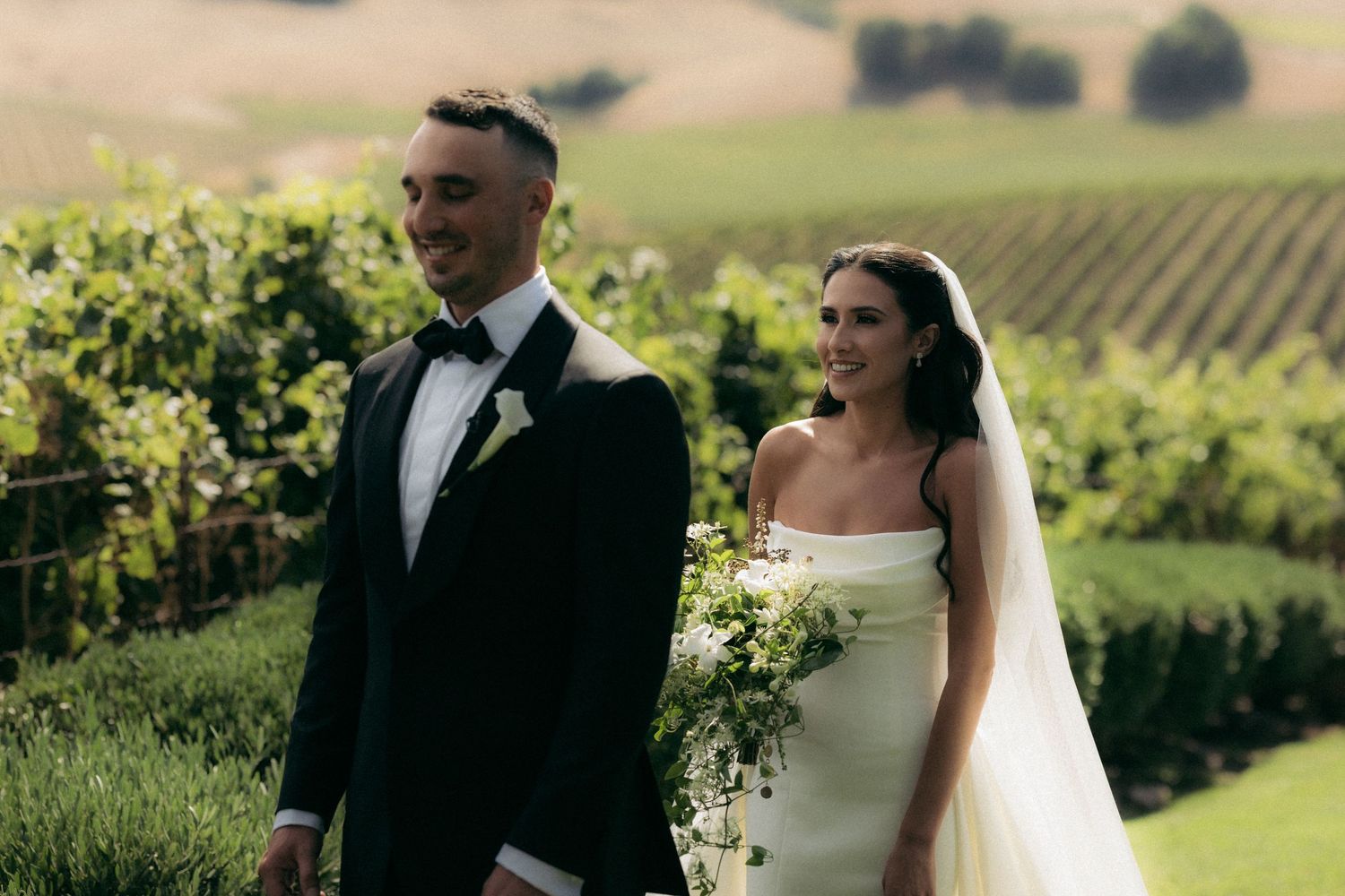 A couple poses together in formal attire against a scenic vineyard backdrop.