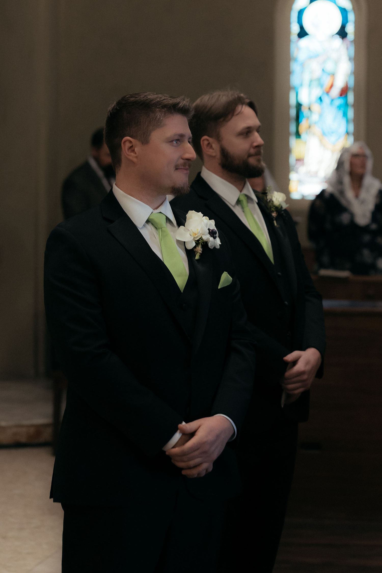 Two groomsmen in black suits with lime green ties stand at attention in a church.