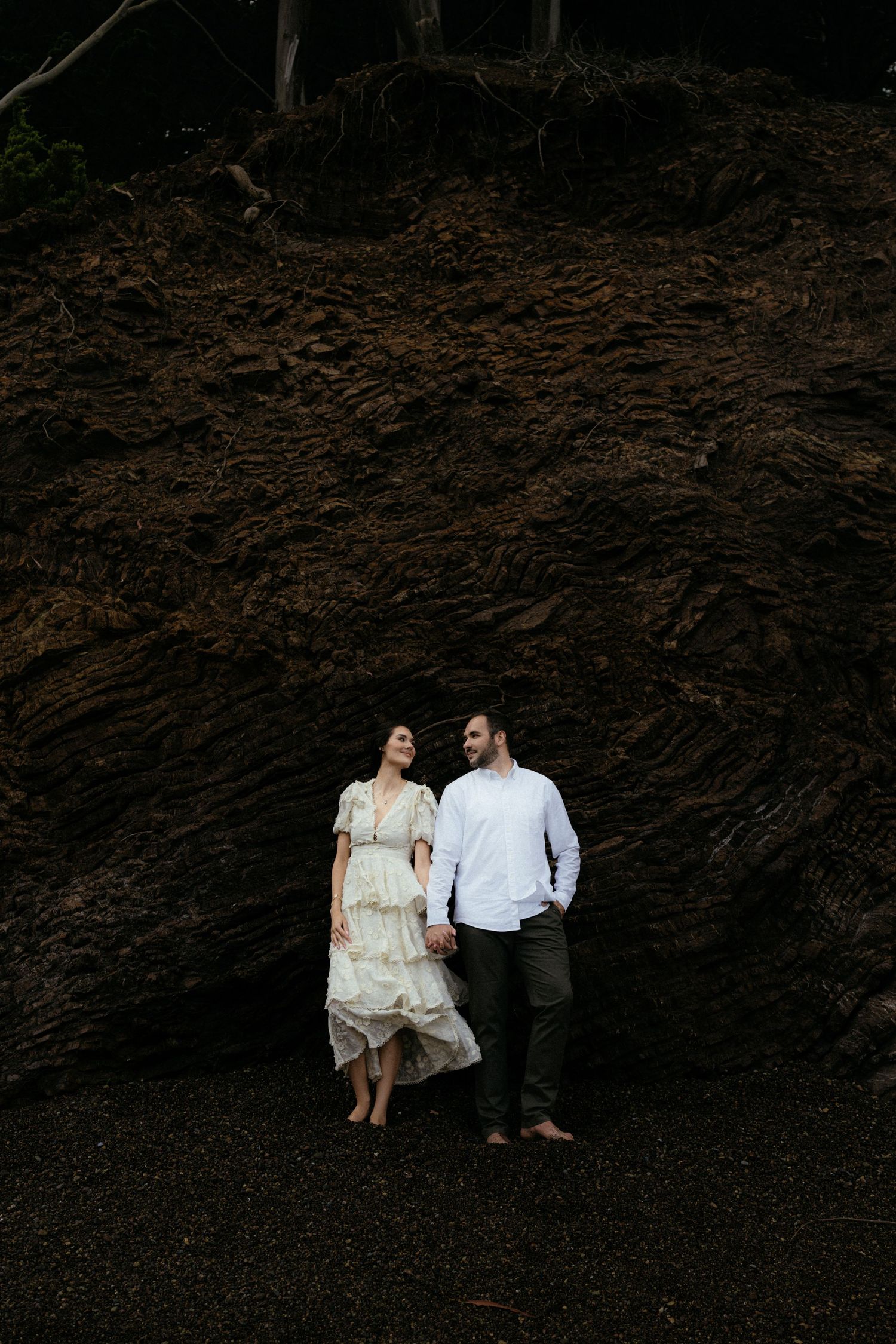 Couple holding hands in front of a large dark rock formation on a beach, woman in white dress, man in white shirt.