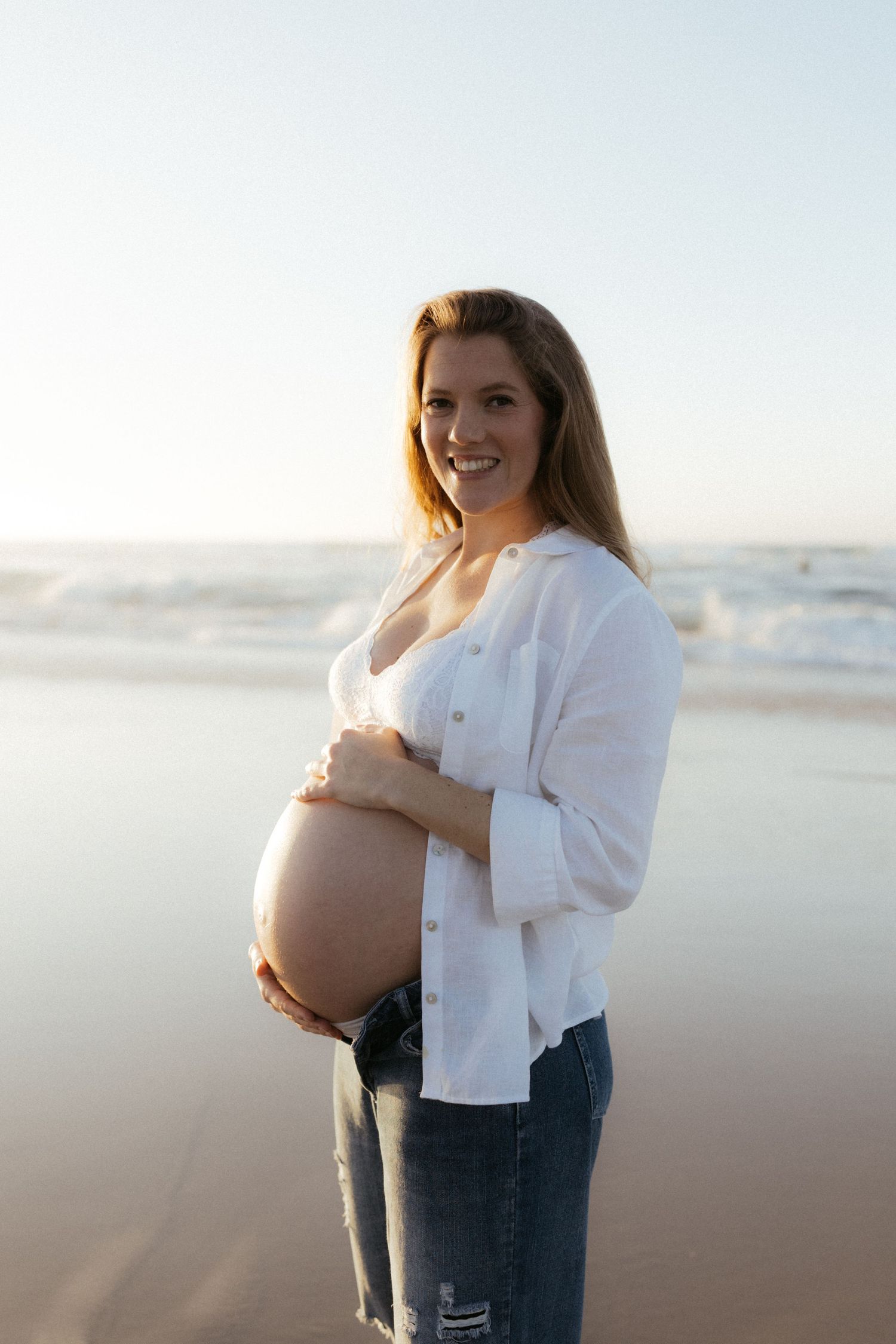 Smiling pregnant woman in white shirt and jeans cradling her baby bump on a sunny beach with ocean waves.