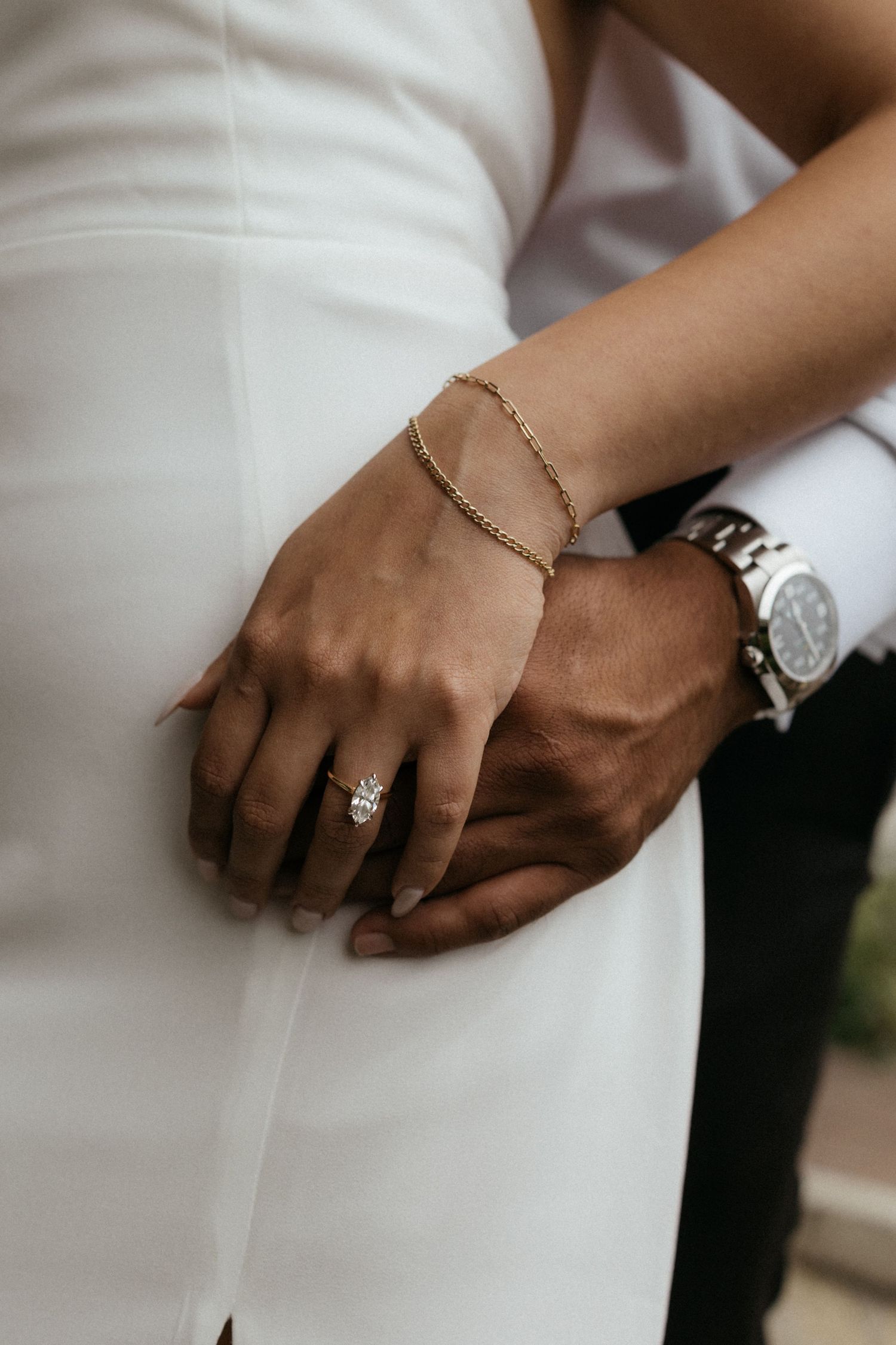 Close-up of bride and groom holding hands, featuring a diamond engagement ring, gold bracelet, and silver watch.