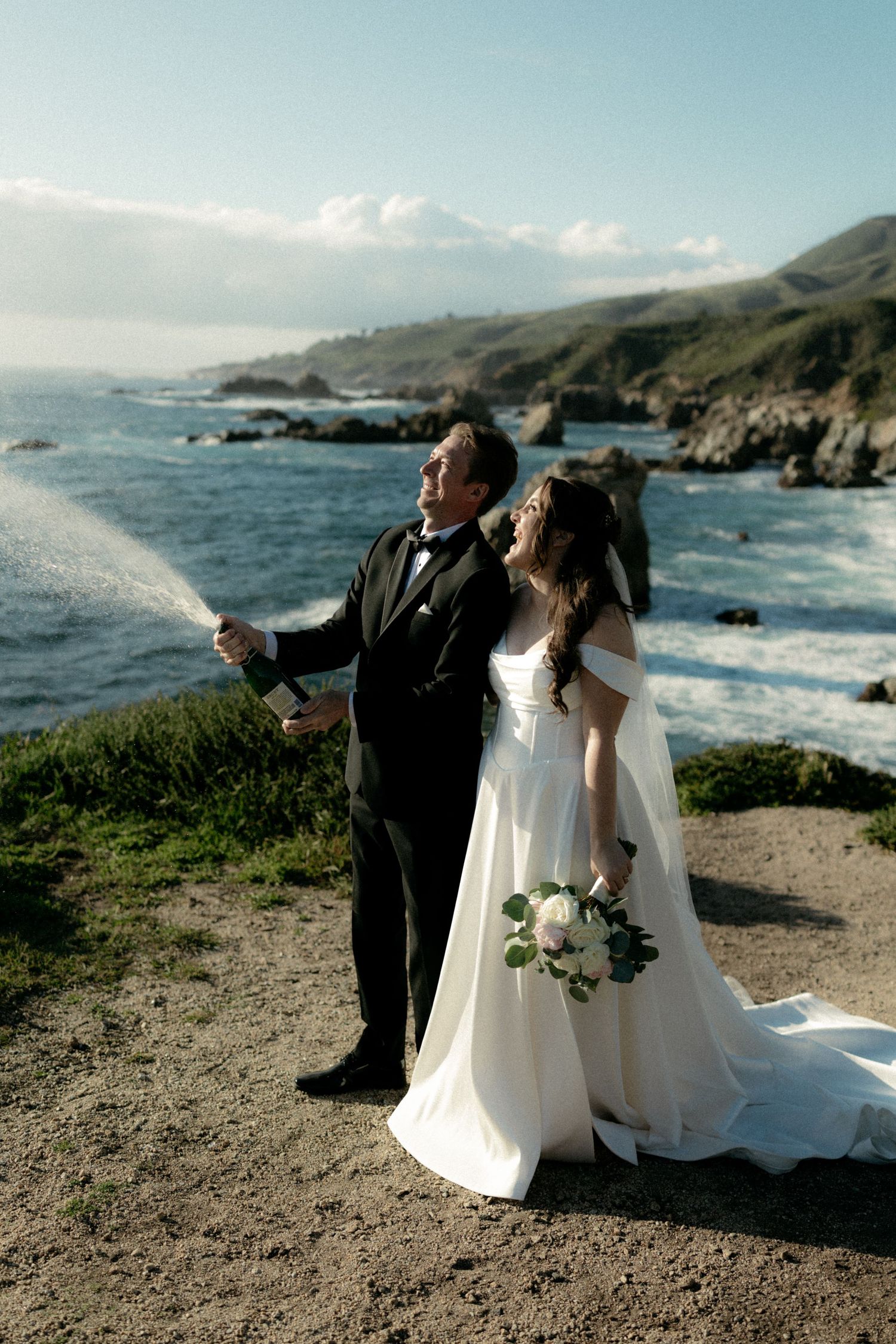 Newlyweds celebrate with champagne spray on coastal cliffside overlooking scenic ocean views.