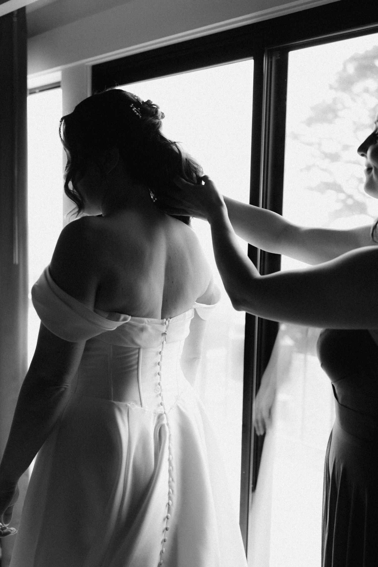 Black and white photo of bride in off-shoulder gown with button back, assisted by bridesmaid near window.