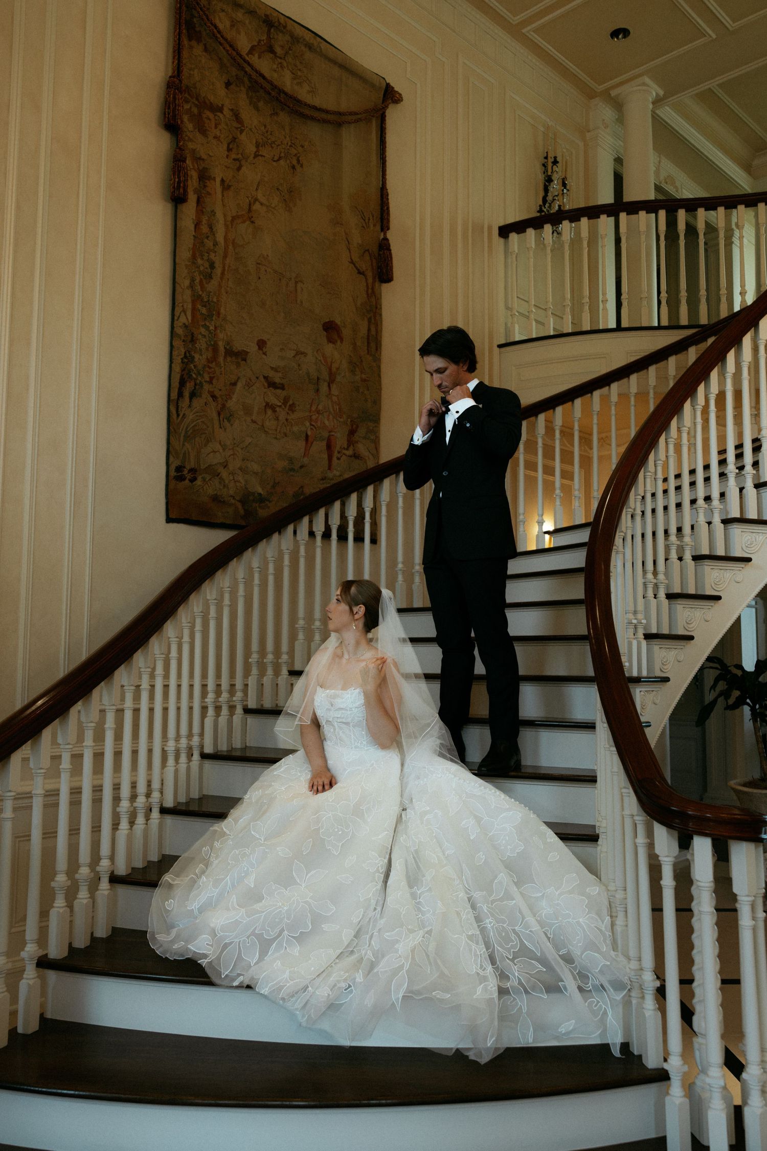 Bride and groom pose on elegant curved staircase, bride in full ball gown with veil, groom in black tuxedo.