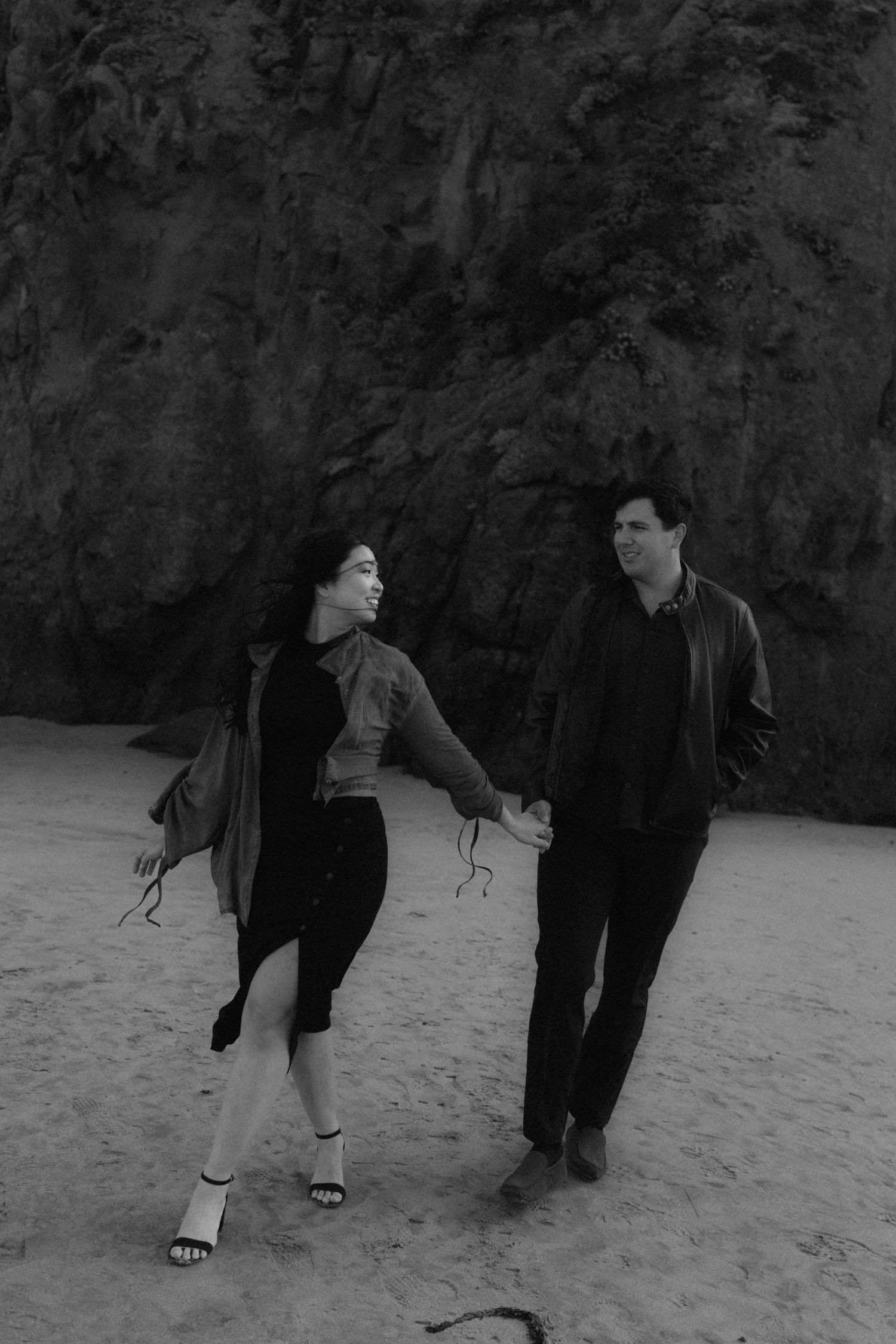 Black and white photo of a couple holding hands and walking on a sandy beach with rocky cliffs behind them.