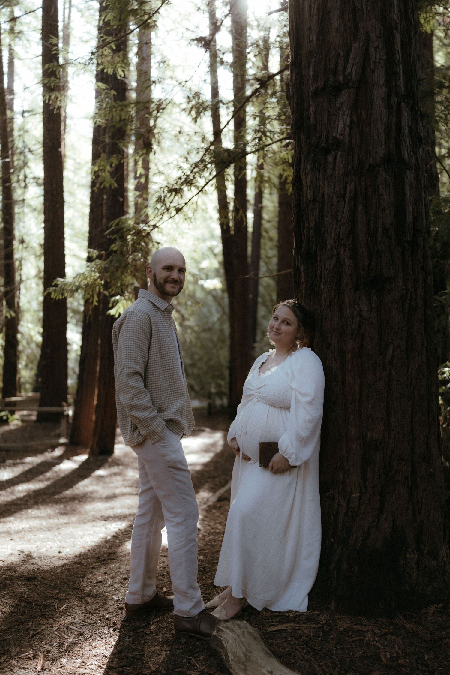 A couple in white attire shares a romantic moment among tall redwood trees.