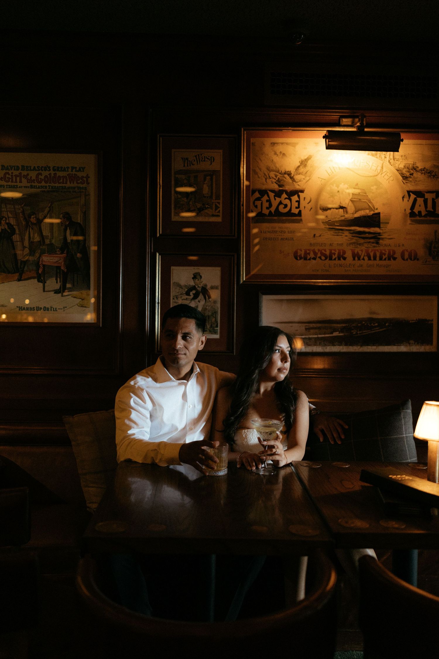 Couple sitting together in a dimly lit vintage bar booth with antique posters and warm ambient lighting.