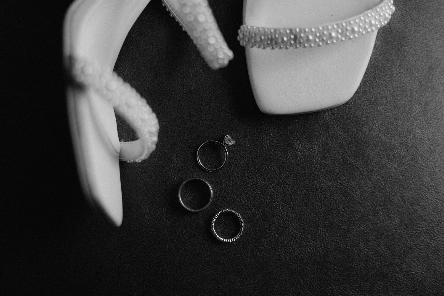 Black and white flat lay of three wedding rings beside white bridal heels with pearl trim on dark background.