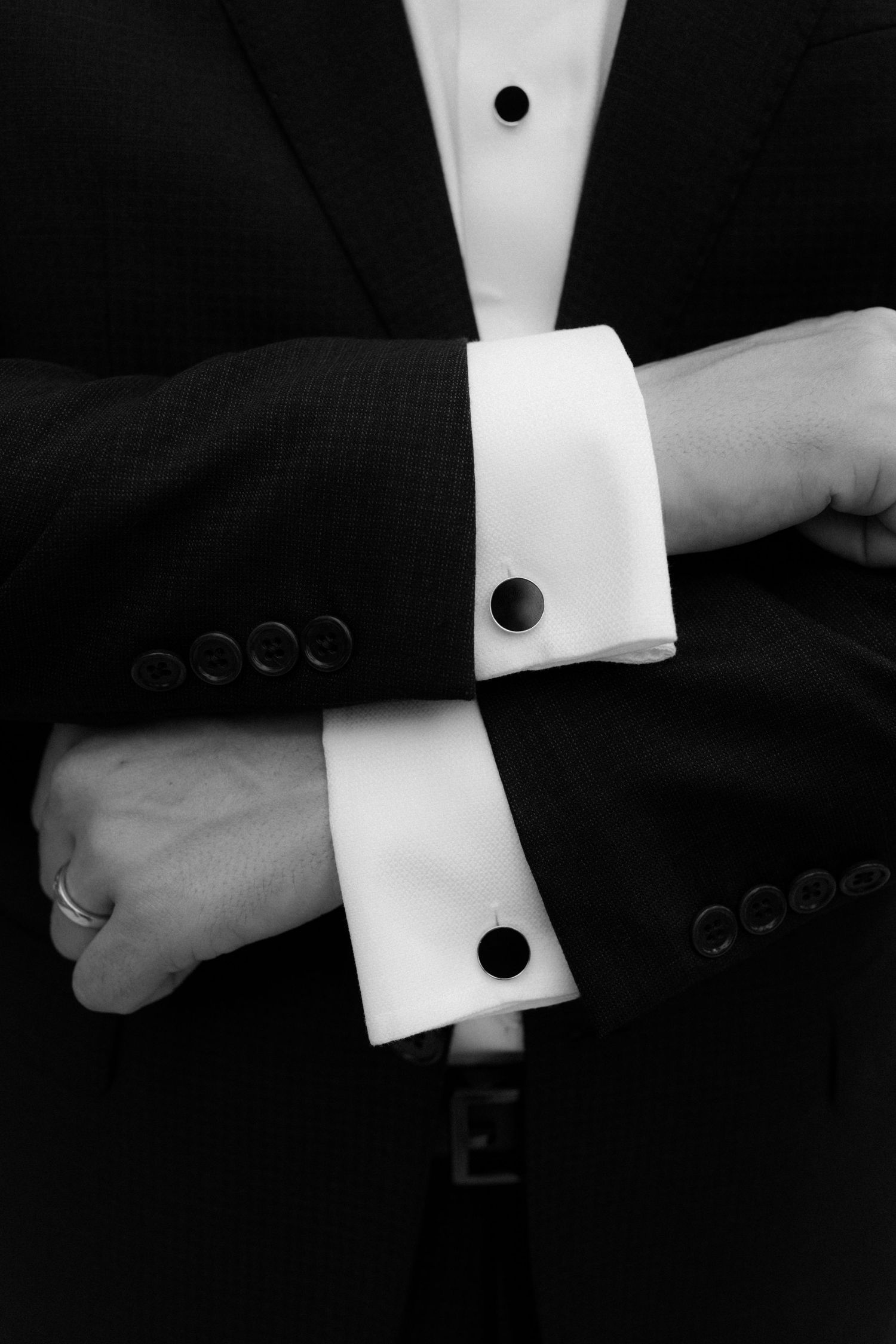 Black and white photo of a man in a tuxedo with white French cuffs and round black cufflinks, arms crossed.