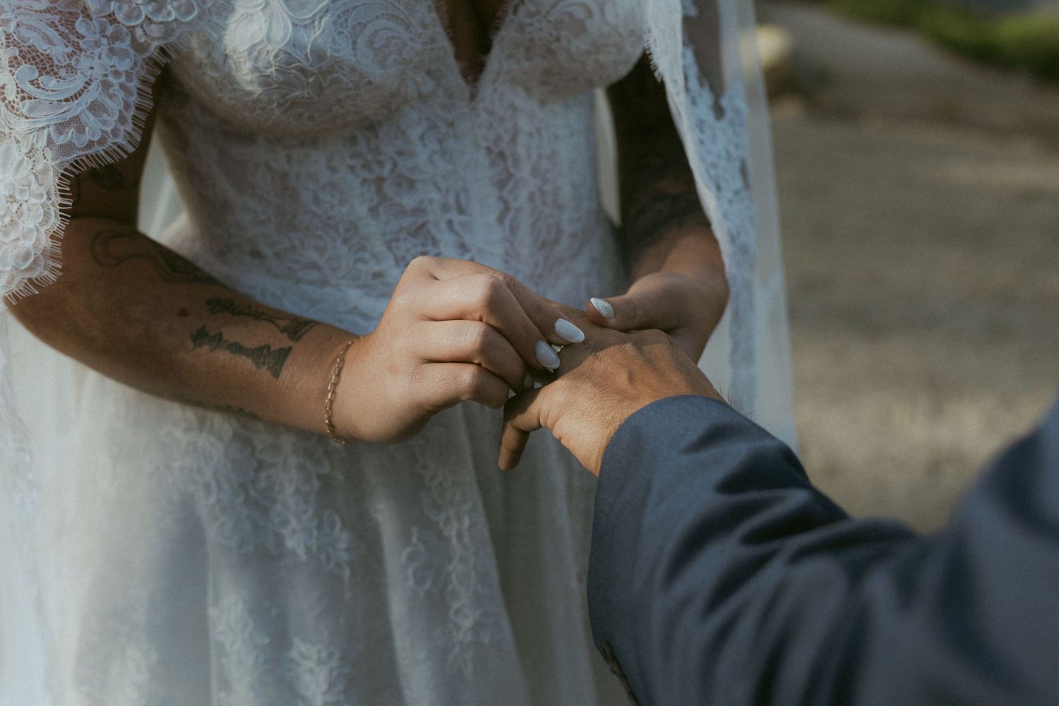 Groom places a wedding ring on the bride's finger, showcasing her tattooed wrist and lace wedding gown.