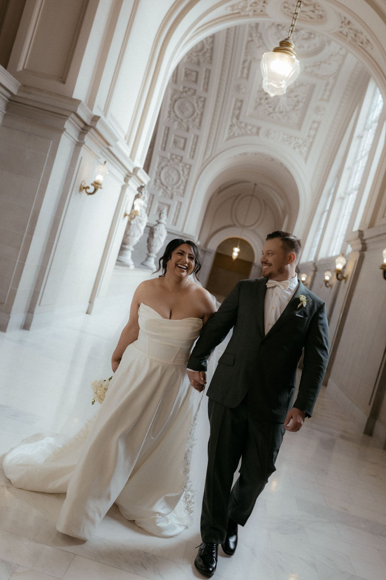 A couple walks through an elegant white hallway with ornate architectural details and arches during their wedding.