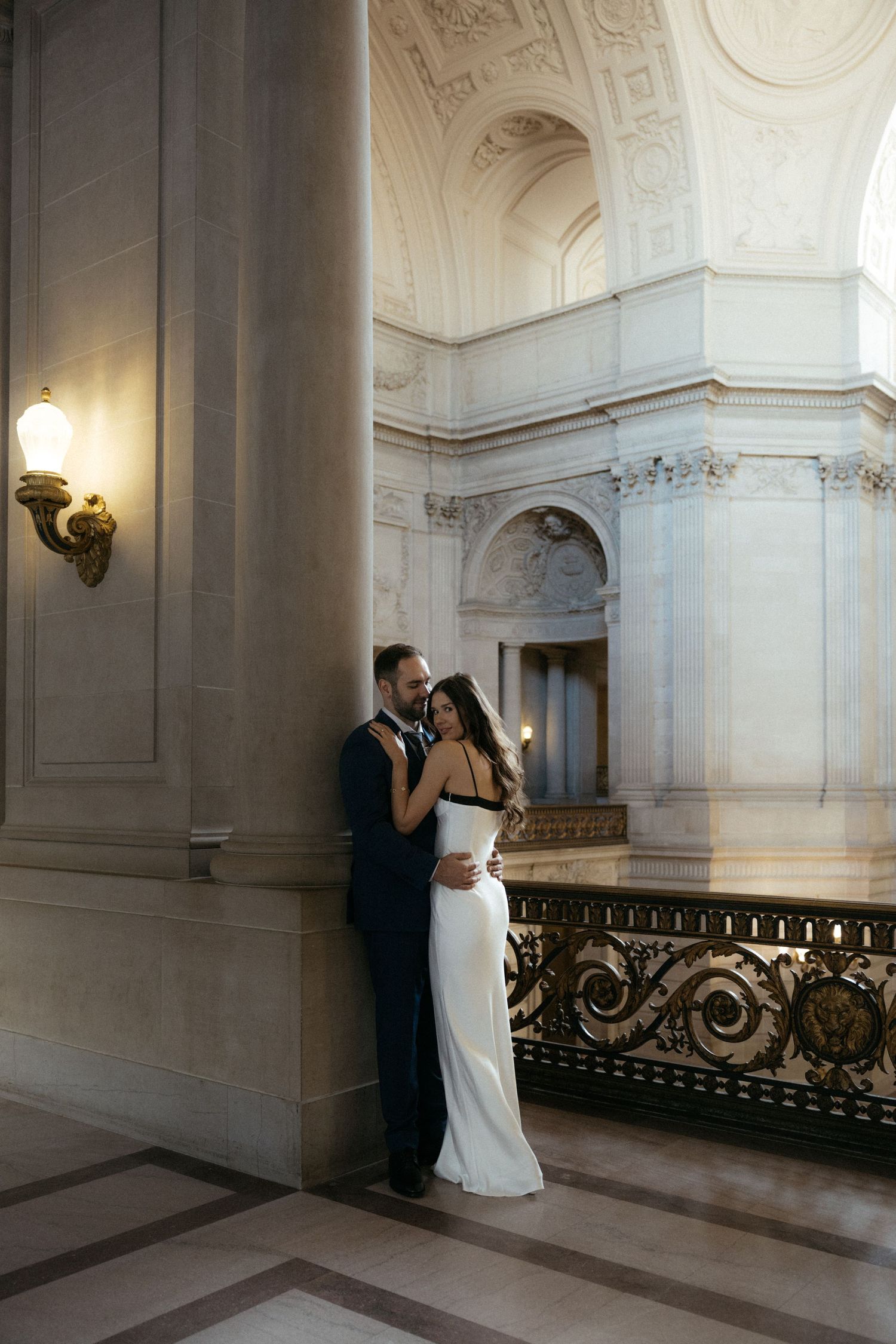 Elegant wedding couple shares romantic moments in a grand marble hallway with ornate architectural details and soft lighting.