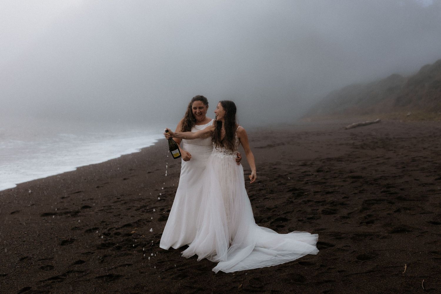 Two brides in flowing white wedding dresses embrace on a misty black sand beach with mountains in the background.