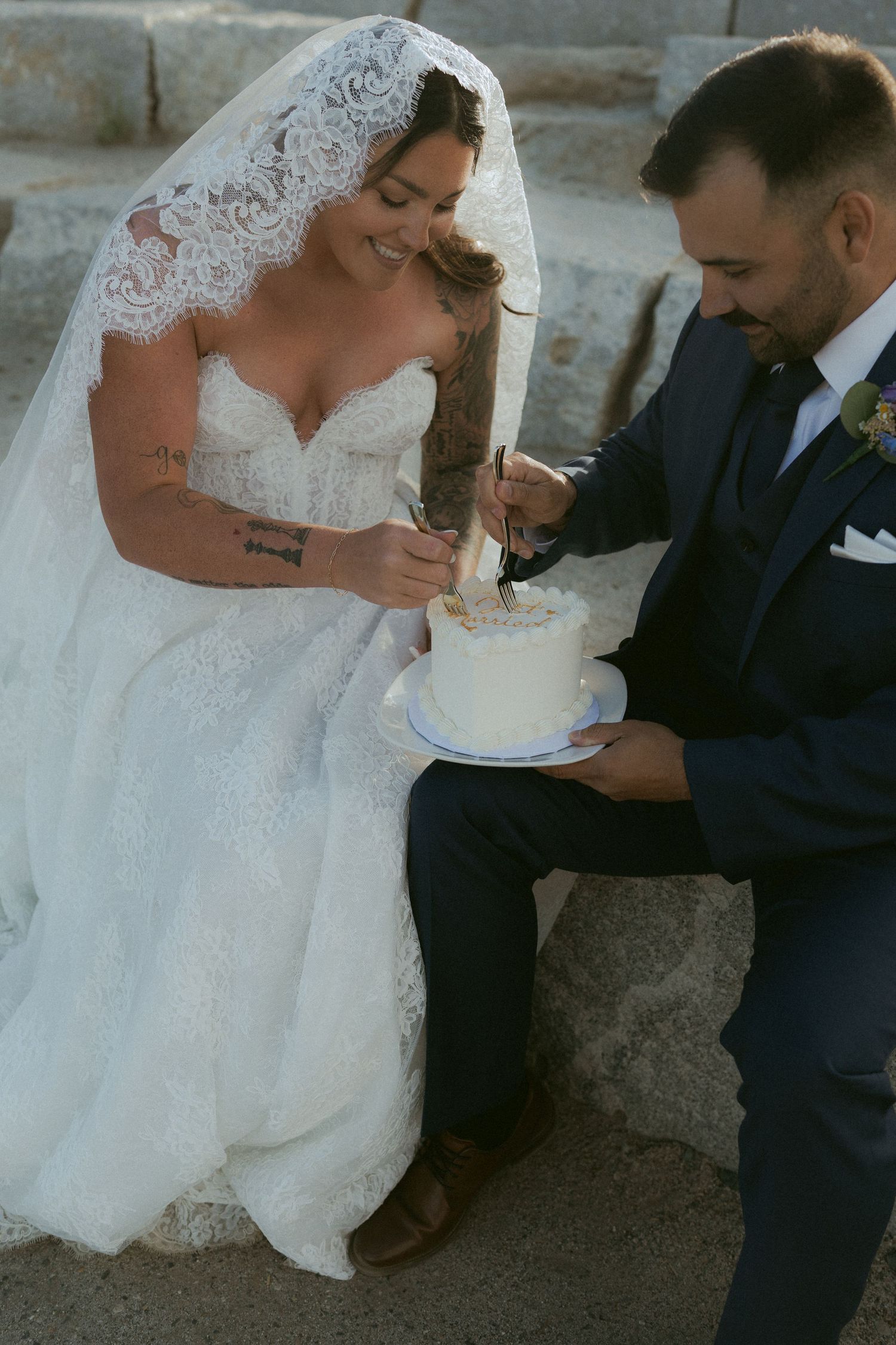 Bride and groom cutting a small white wedding cake together outdoors, bride wearing lace gown and veil.