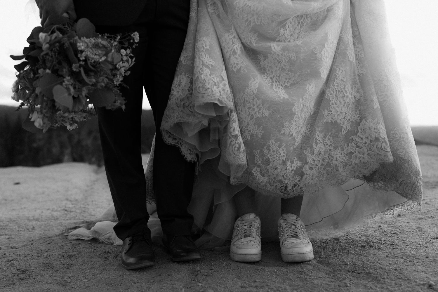 Black and white wedding photo showing bride in lace gown with sneakers and groom holding bouquet, standing on sandy ground.