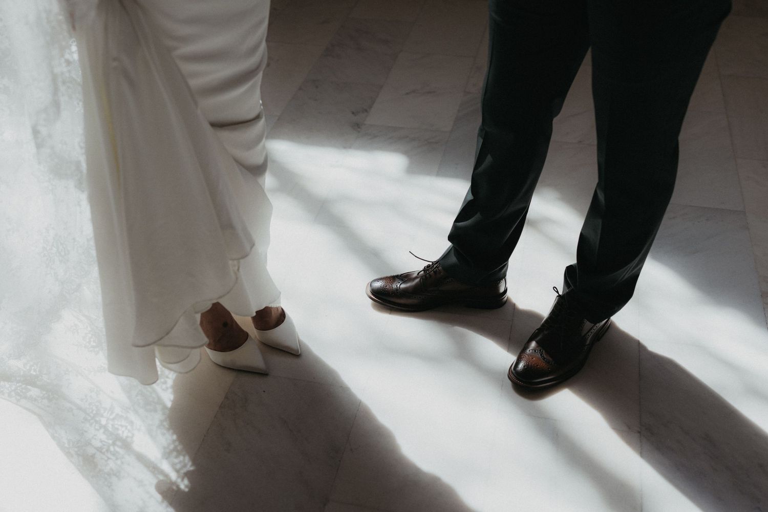 Wedding shoes and formal attire captured in dramatic lighting on a marble floor.