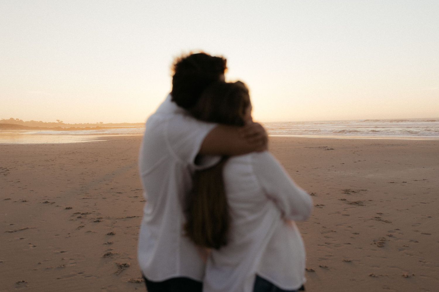 Couple embracing on a sandy beach at sunset, wearing white outfits with ocean waves in the background.