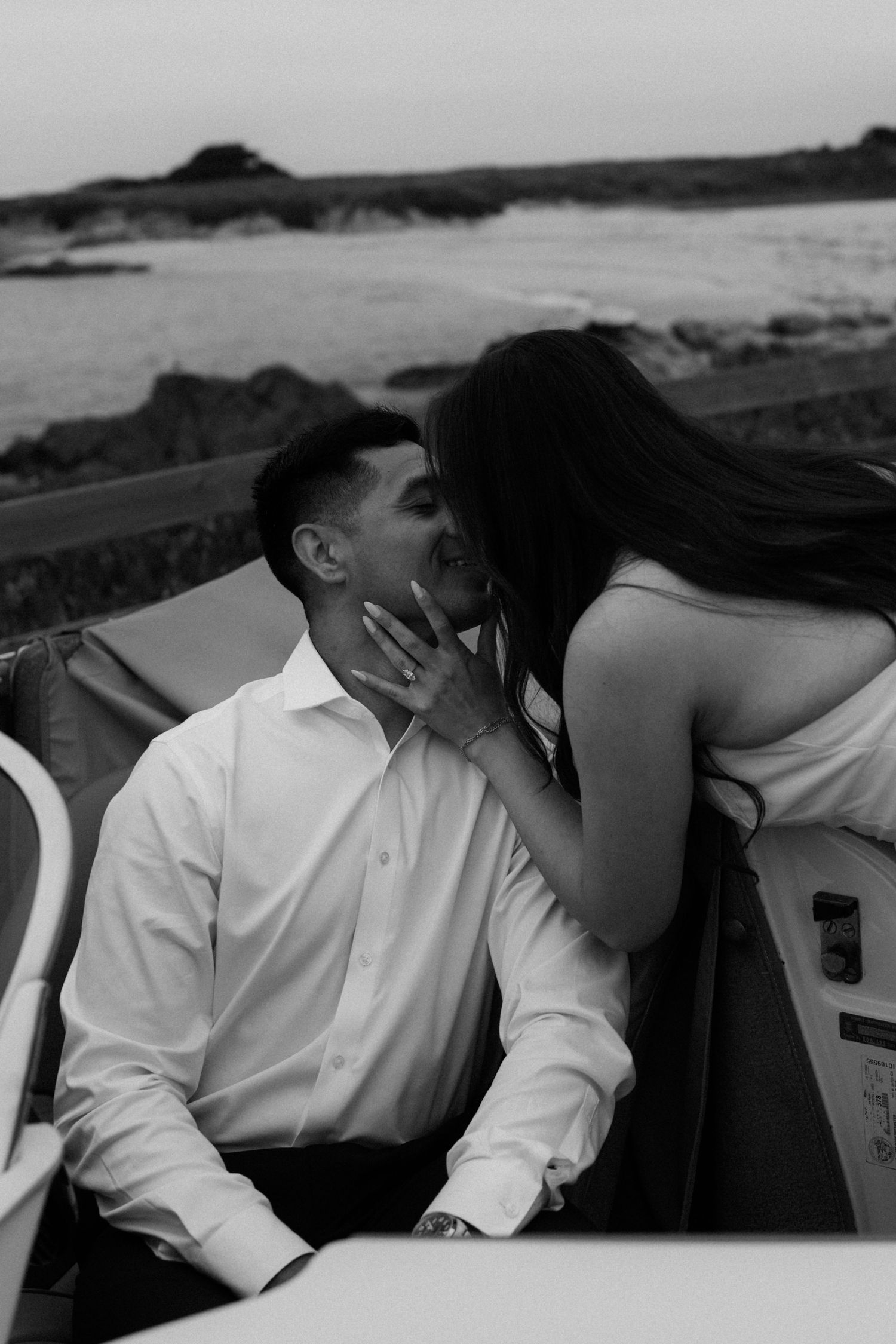 Black and white photo of a couple sharing a romantic kiss on a boat near a rocky coastline at dusk.