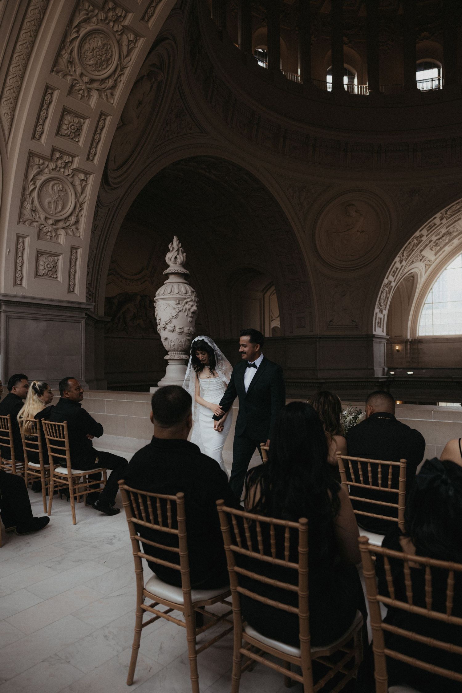 Bride and groom holding hands at wedding ceremony inside ornate San Francisco City Hall with gold chairs and guests.