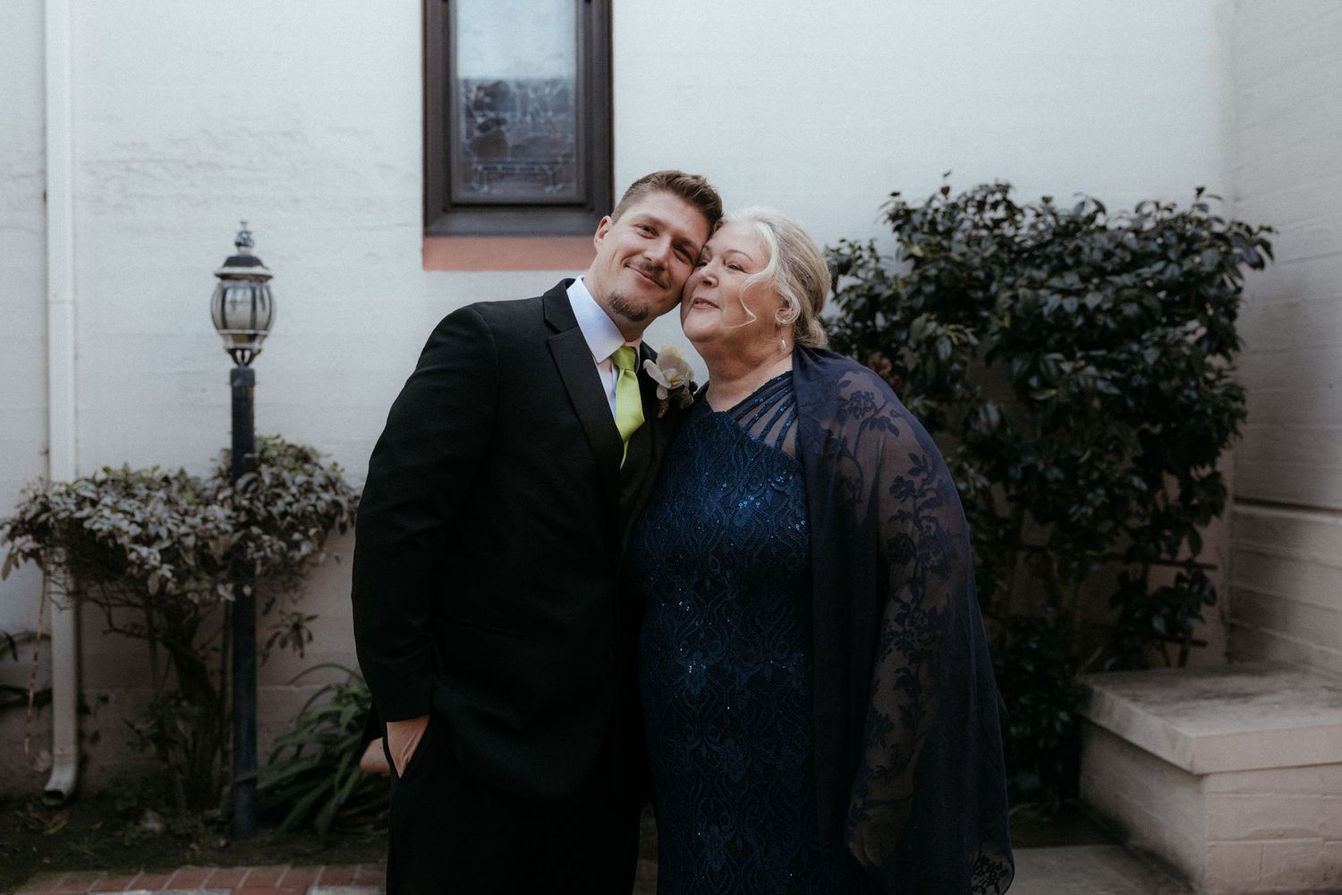 A couple shares an intimate kiss while standing against a white exterior wall near greenery and outdoor lighting.